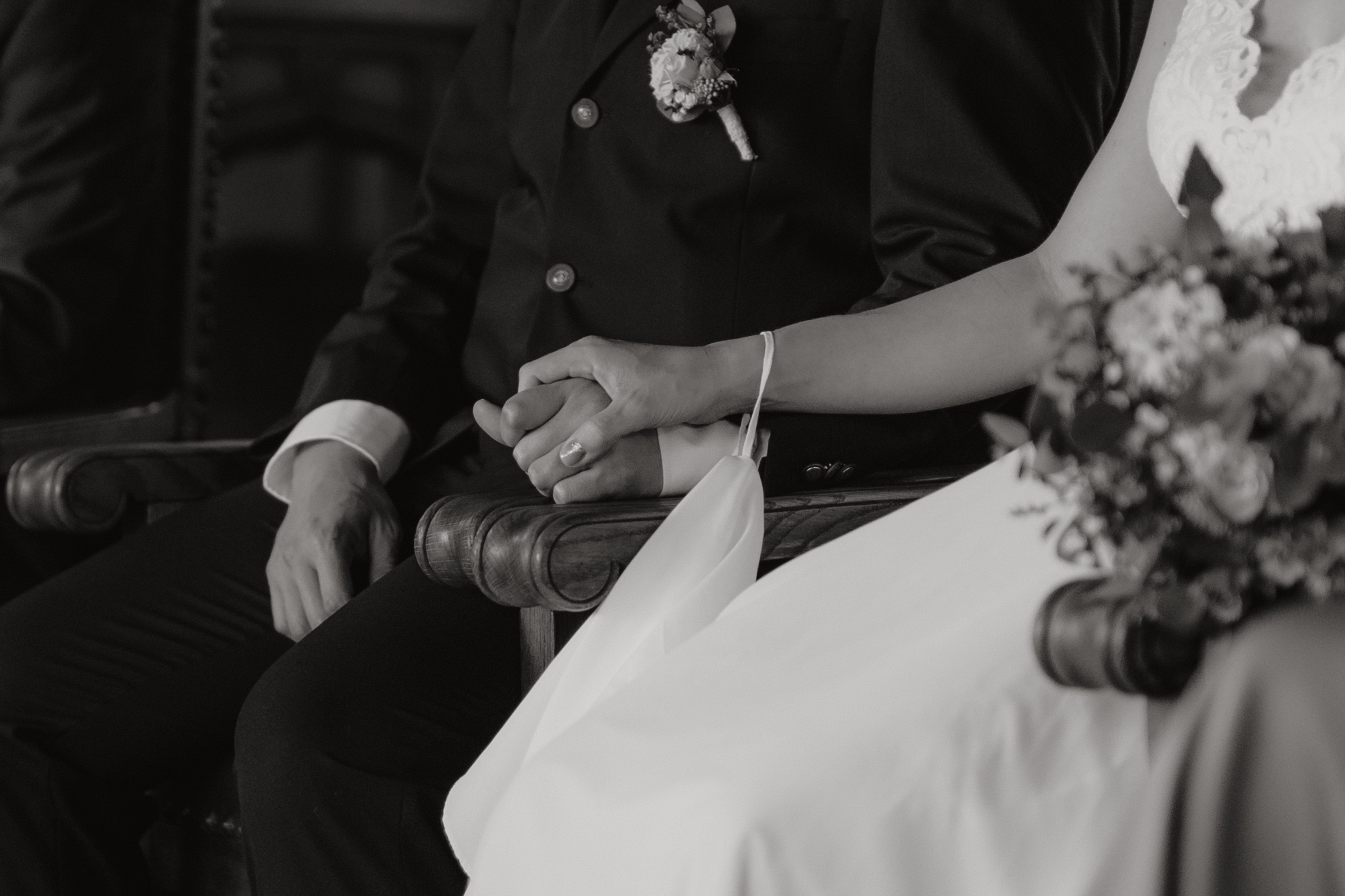 Eine Hochzeitsfotografie von Tasha Ender Photography zeigt eine Nahaufnahme eines Paares, das Seite an Seite sitzt und Händchen hält. Die Frau trägt ein weißes Hochzeitskleid mit einem Blumenstrauß auf ihrem Schoß, während der Mann einen dunklen Anzug mit einer Boutonniere an seiner Jacke trägt und ihre Zivilhochzeit in Baden feiert.