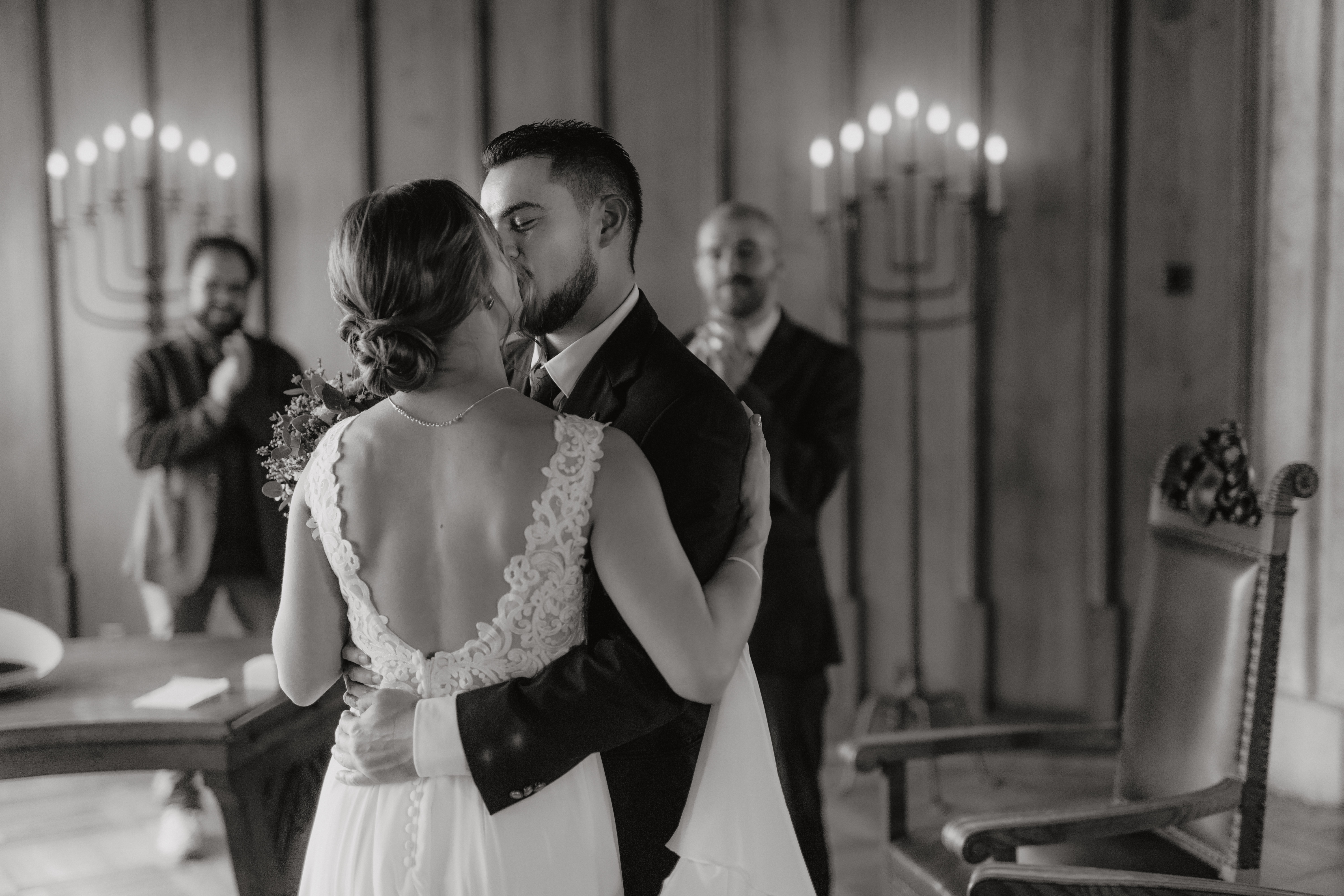 Während ihrer Zivilhochzeit in Baden küssen sich Braut und Bräutigam. Das weiße Kleid der Braut, das auf dem Rücken mit Spitzendetails verziert ist, bildet einen schönen Kontrast zum eleganten Anzug des Bräutigams. Im Hintergrund schauen zwei Männer neben hohen Kerzenständern liebevoll zu. Dieser ergreifende Moment im Aargau wurde von Hochzeitsfotografie von Tasha Ender Photography in Schwarzweiß wunderschön festgehalten.