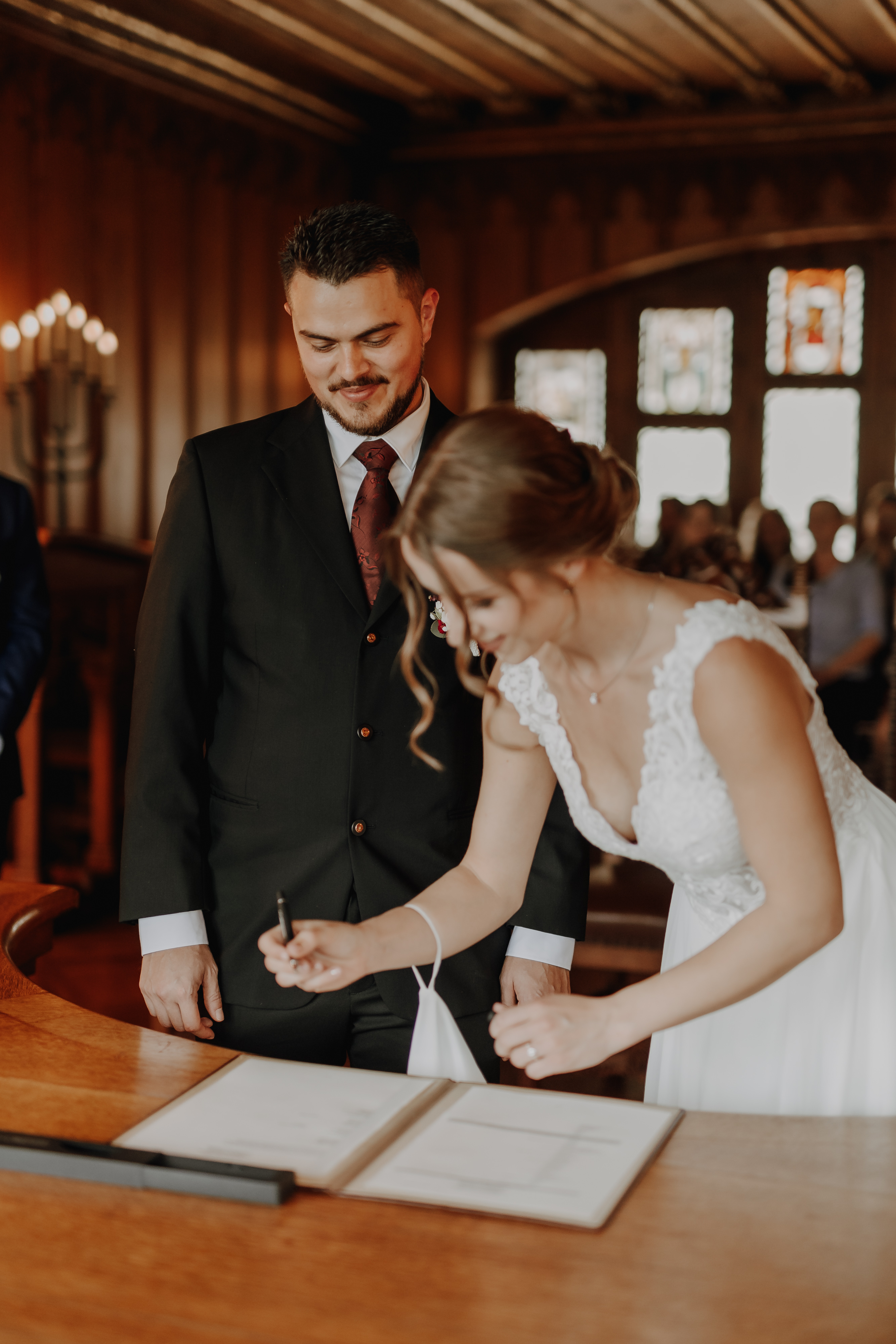 Eine Braut im weißen Kleid unterschreibt ein Hochzeitsdokument auf einem Holztisch, während der Bräutigam, in einen schwarzen Anzug gekleidet, sie lächelnd beobachtet. Sie befinden sich in einem warm beleuchteten, rustikalen Raum mit Holzwänden und Buntglasfenstern. Im Hintergrund sind die Gäste verschwommen, die ihre Ziviltrauung im Aargau feiern. Dieser besondere Moment wurde von Tasha Ender Photography's Hochzeitsfotografie wunderschön festgehalten.