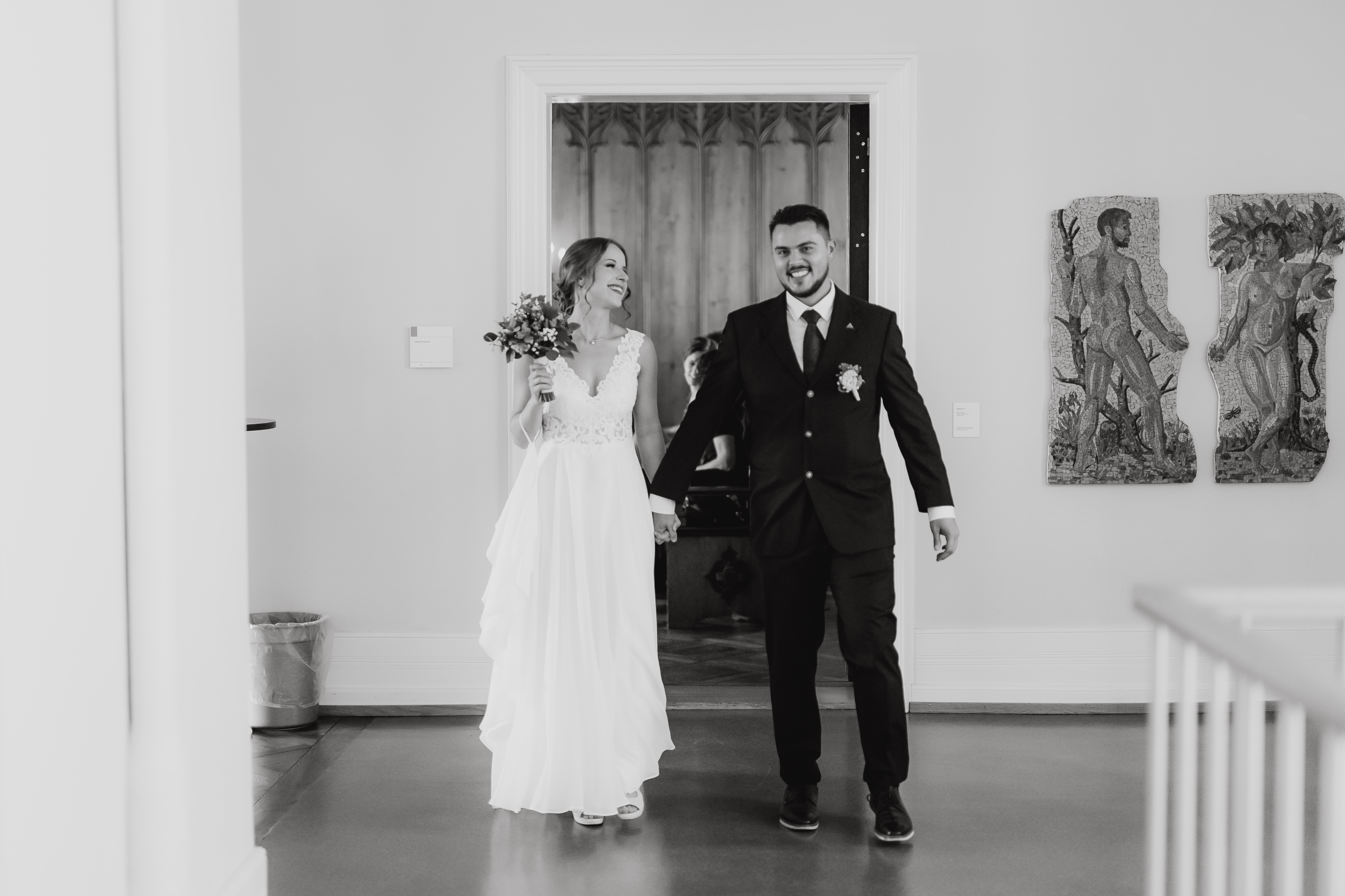 Eine lächelnde Braut in einem fließenden weißen Kleid hält einen Blumenstrauß und geht Hand in Hand mit einem Bräutigam in schwarzem Anzug und Krawatte. Sie sind in einem Haus in Aargau für ihre Ziviltrauung und an der Wand hinter ihnen hängen zwei Kunstwerke mit menschlichen Figuren. Die Szene strahlt Freude und Feierlichkeit aus während dieser besonderen Hochzeitsfotografie-Sitzung von Tasha Ender Photography.