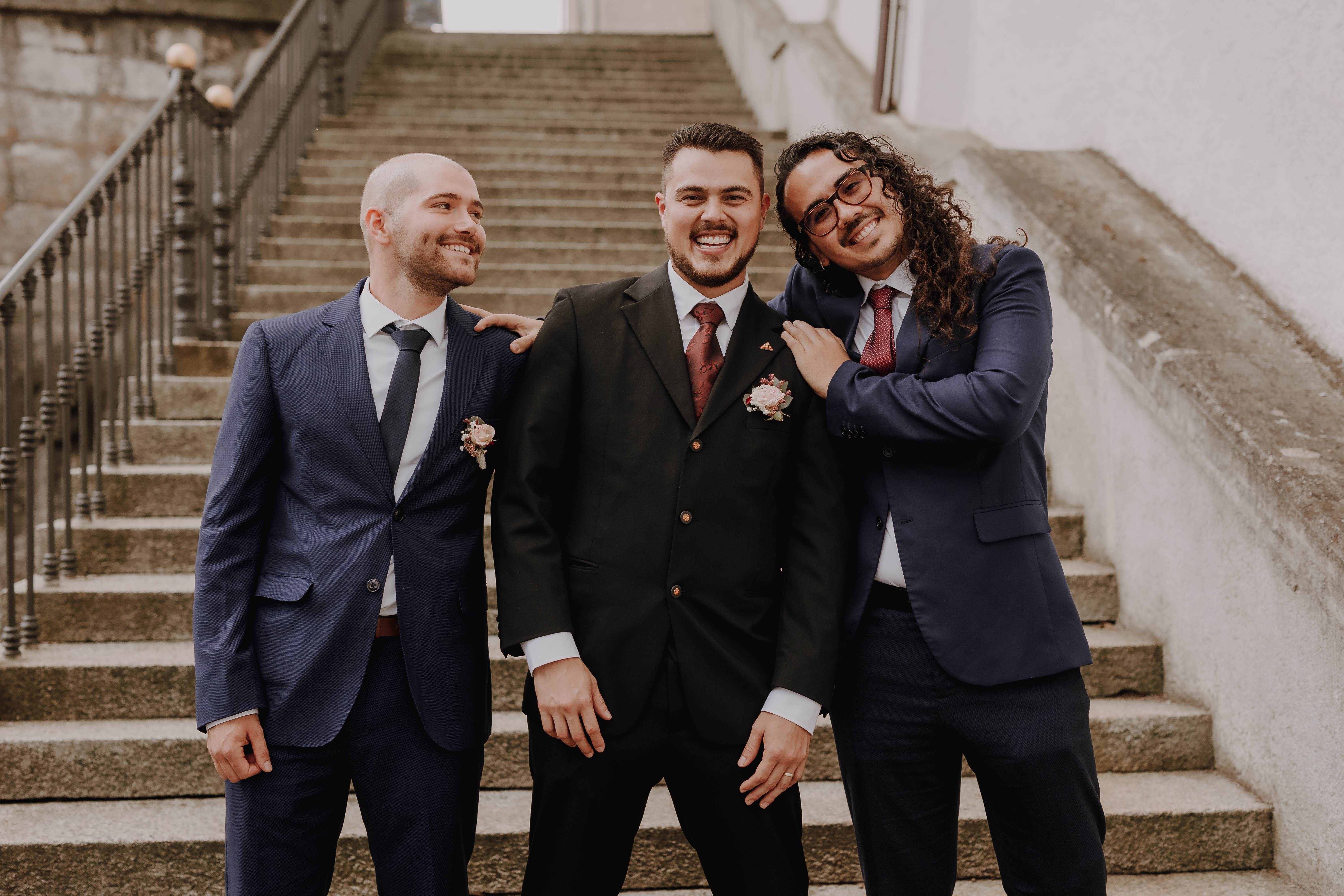 Drei Männer posieren gemeinsam vor einer Treppe im Aargau. Der Mann in der Mitte, elegant gekleidet in einem schwarzen Anzug, lächelt breit und lässt seine Hände entspannt an den Seiten hängen. Die beiden Männer neben ihm, beide in blauen Anzügen und feiern ihre Ziviltrauung, stehen dicht beieinander und legen liebevoll ihre Hände auf seine Schultern. Dieser berührende Moment wurde vom Hochzeitsfotografie-Service von Tasha Ender Photography wunderschön festgehalten.