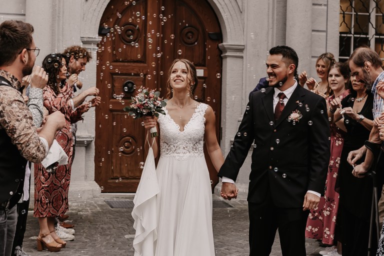 Braut und Bräutigam halten sich an den Händen und lächeln fröhlich, als sie ein Gebäude in Baden verlassen, umgeben von Freunden und Familie, die Seifenblasen machen. Die Braut, Tasha Ender, hält einen Blumenstrauß in der Hand und beide tragen Hochzeitskleidung.