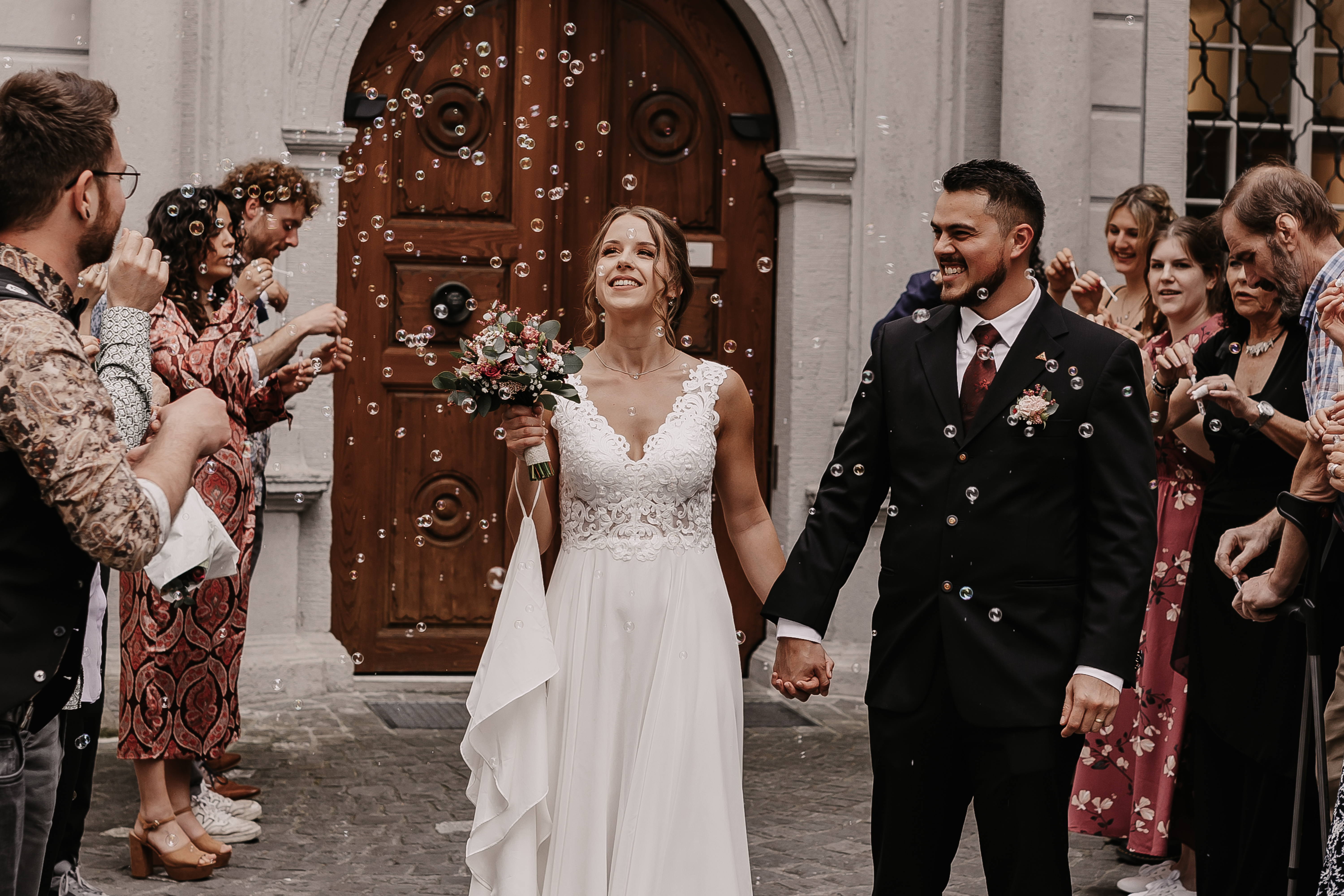 Braut und Bräutigam halten sich an den Händen und lächeln fröhlich, als sie ein Gebäude in Baden verlassen, umgeben von Freunden und Familie, die Seifenblasen machen. Die Braut, Tasha Ender, hält einen Blumenstrauß in der Hand und beide tragen Hochzeitskleidung.
