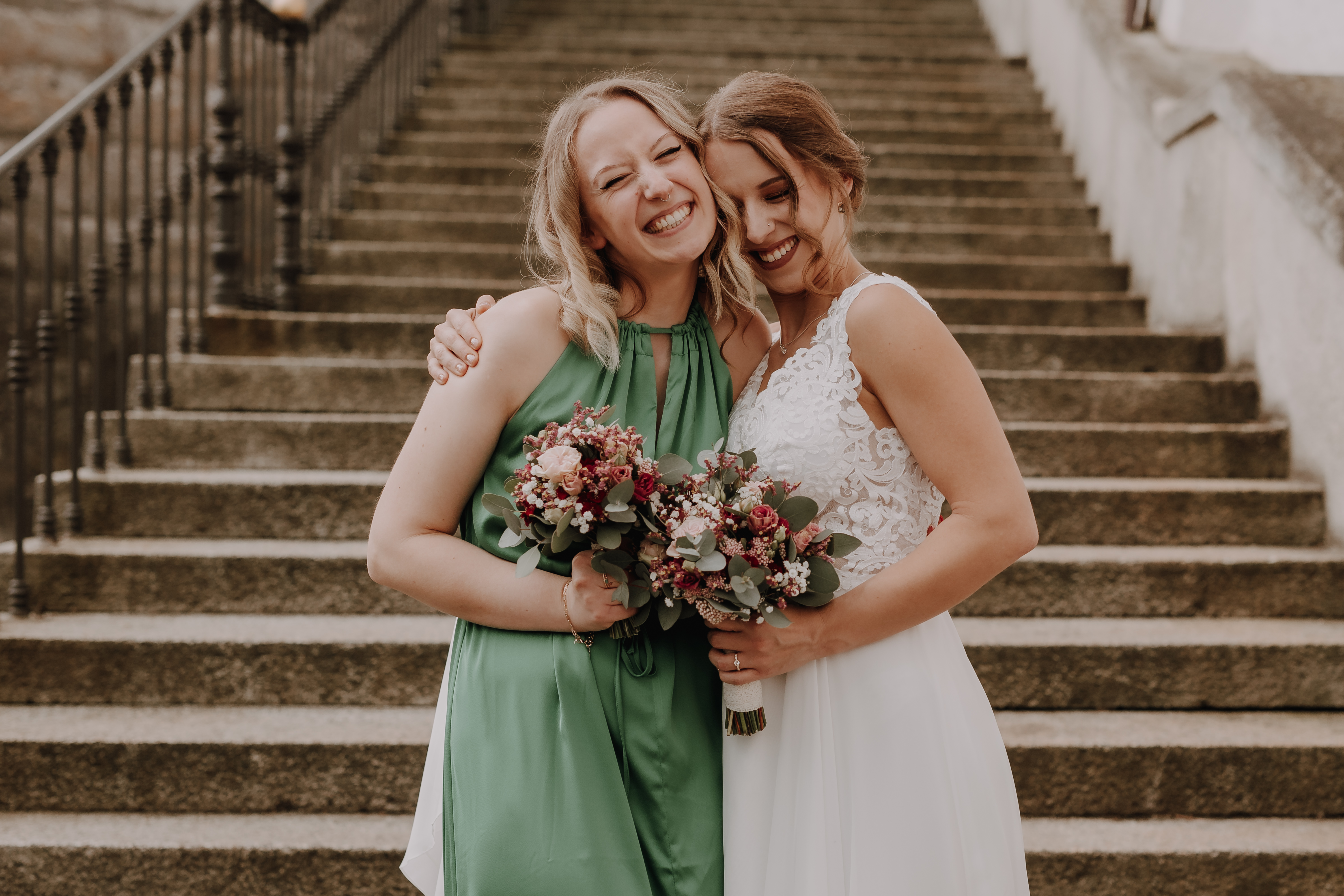Zwei Frauen, die erste in einem weißen Brautkleid und die zweite in einem grünen Kleid, stehen auf Steinstufen in Baden. Beide lächeln und halten Blumensträuße in den Händen. Sie umarmen sich freudig und feiern wahrscheinlich eine Zivilhochzeit oder einen anderen besonderen Anlass. Dieser herzerwärmende Moment wurde vom Hochzeitsfotografie-Service von Tasha Ender Photography wunderschön festgehalten.