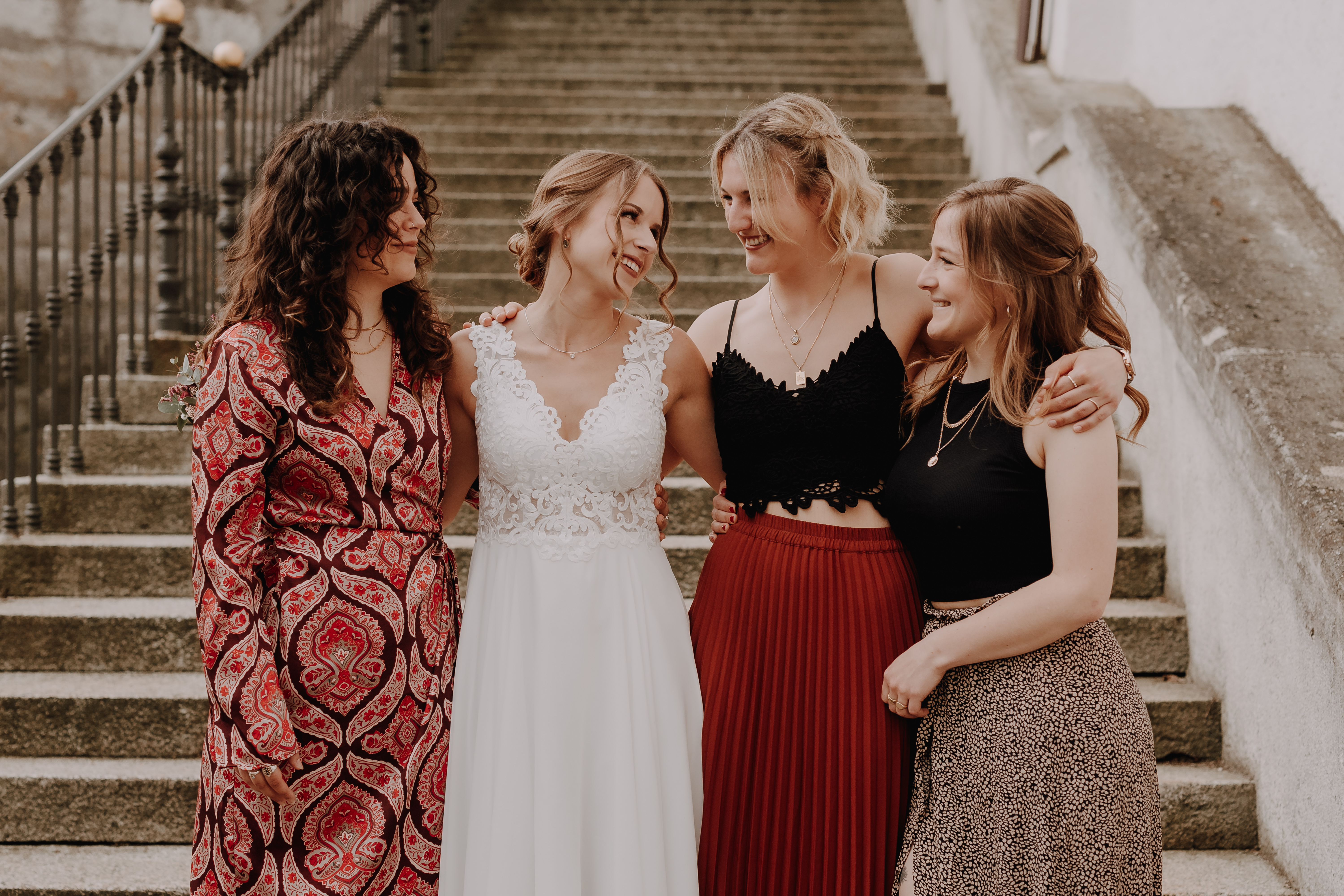 Vier Frauen stehen auf einer Treppe in Baden, lächeln und haben die Arme umeinander gelegt. Die Frau links trägt ein rot gemustertes Kleid, die Frau neben ihr ein weißes Kleid. Neben ihr trägt eine andere ein schwarzes Oberteil mit einem roten Rock und die vierte Frau trägt ein schwarzes Oberteil mit einem gemusterten Rock. Ihre Hochzeitsfotografie-Session von Tasha Ender Photography fängt echte Freude und Stil ein, akzentuiert durch