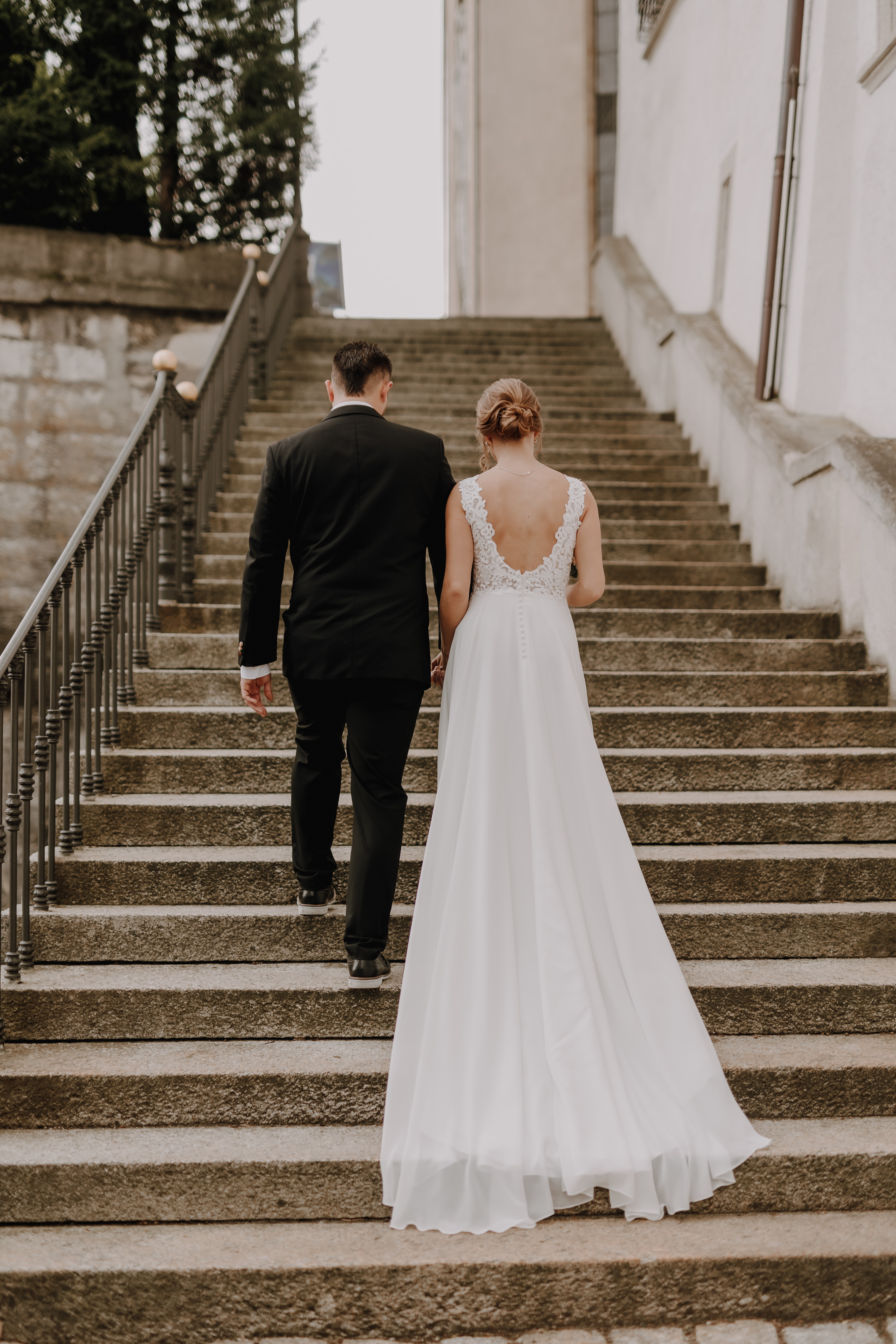 Ein Paar, von hinten gesehen, geht eine Steintreppe hinauf. Die Frau trägt ein langes, ärmelloses weißes Kleid mit Spitzenrücken und der Mann einen schwarzen Anzug. Dieser heitere Moment fängt ihre Ziviltrauung in Aargau ein, mit den Stufen, die von Steinmauern und Metallgeländern inmitten üppigen Grüns gesäumt sind. Aufgenommen von Tasha Ender Photography im Rahmen ihres Hochzeitsfotografie-Dienstes, verewigt dieses Foto den intimen Anlass des Paares auf wunderschöne Weise.