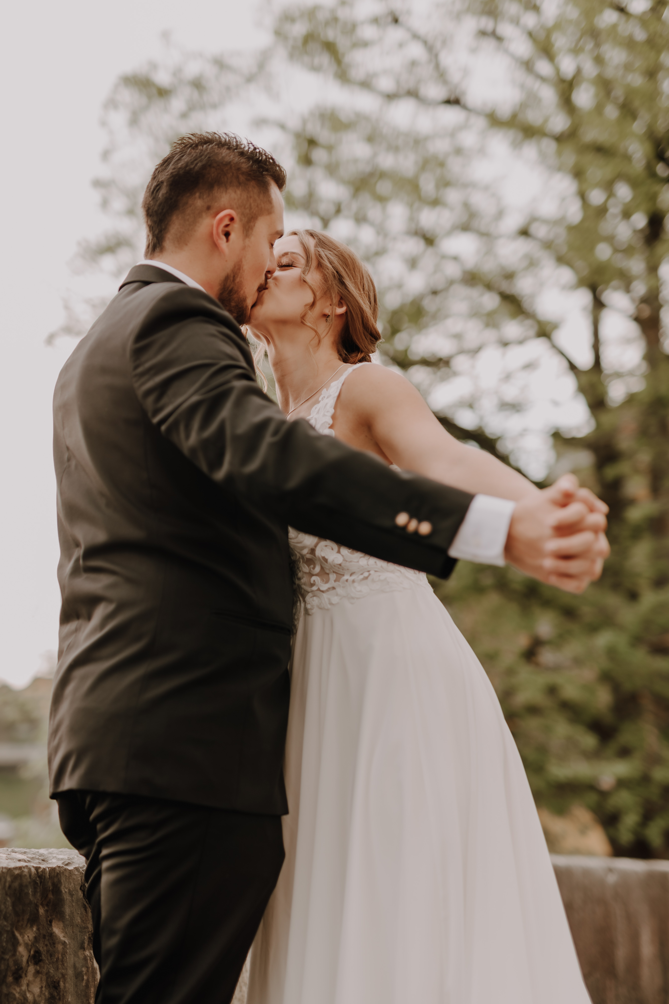 Ein Paar in Hochzeitskleidung küsst sich im Freien während seiner Ziviltrauung. Die Braut hält die Hände des Bräutigams, während sie eng beieinander stehen, umgeben von Bäumen im Hintergrund. Der Bräutigam trägt einen schwarzen Anzug und die Braut ein weißes Hochzeitskleid, perfekt eingefangen von Hochzeitsfotografie.