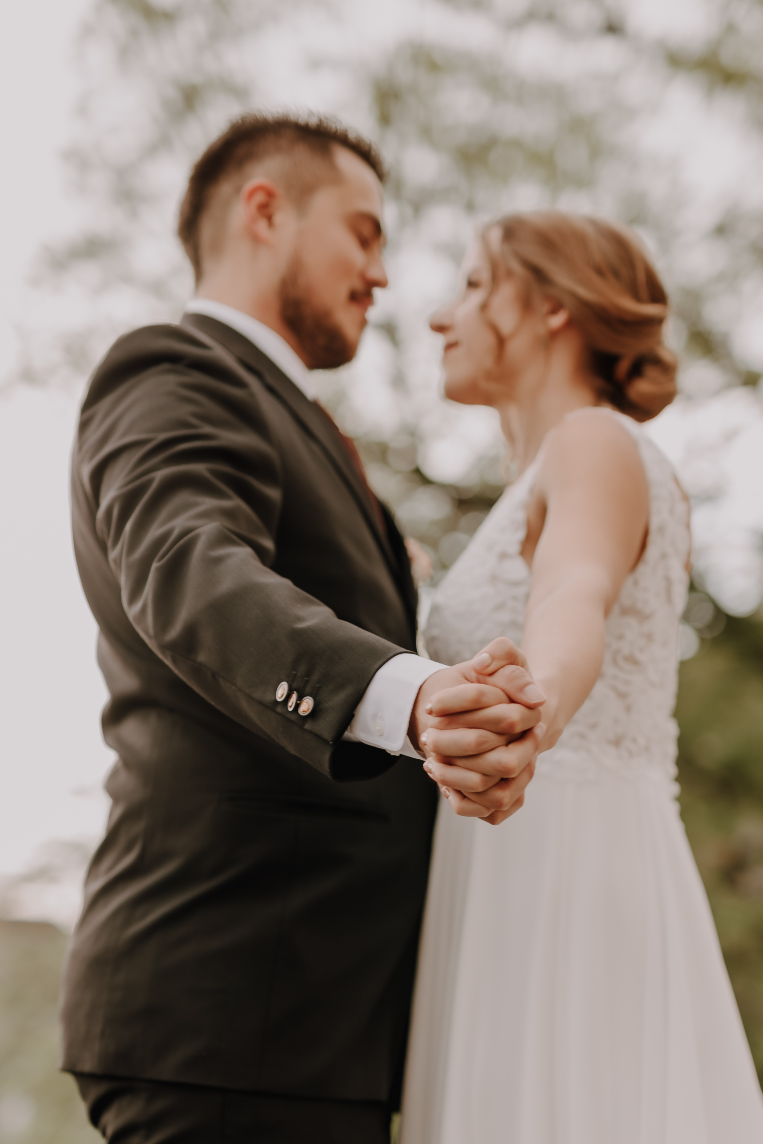 Ein Paar steht eng beieinander, hält sich an den Händen und blickt sich in die Augen. Der Mann trägt einen dunklen Anzug, während die Frau ein weißes ärmelloses Kleid trägt. Die sanft verschwommenen Bäume im Hintergrund erzeugen eine romantische Atmosphäre, die an eine malerische Hochzeit von Tasha Ender erinnert.