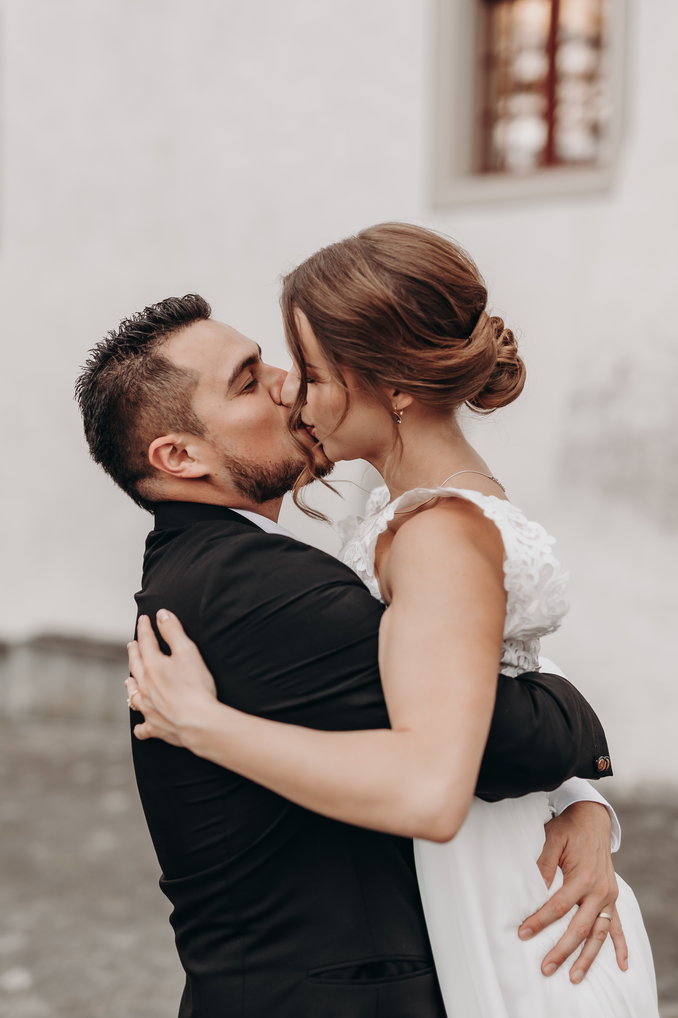 Ein Paar im Hochzeitskleid küsst sich leidenschaftlich. Der Bräutigam, in einen schwarzen Anzug gekleidet, umarmt die Braut, die ein weißes Hochzeitskleid mit Spitzendetails trägt. Sie stehen vor einer cremefarbenen Wand mit einem Fenster und sind in dem Moment verloren – eine perfekte Aufnahme für Hochzeitsfotografie.