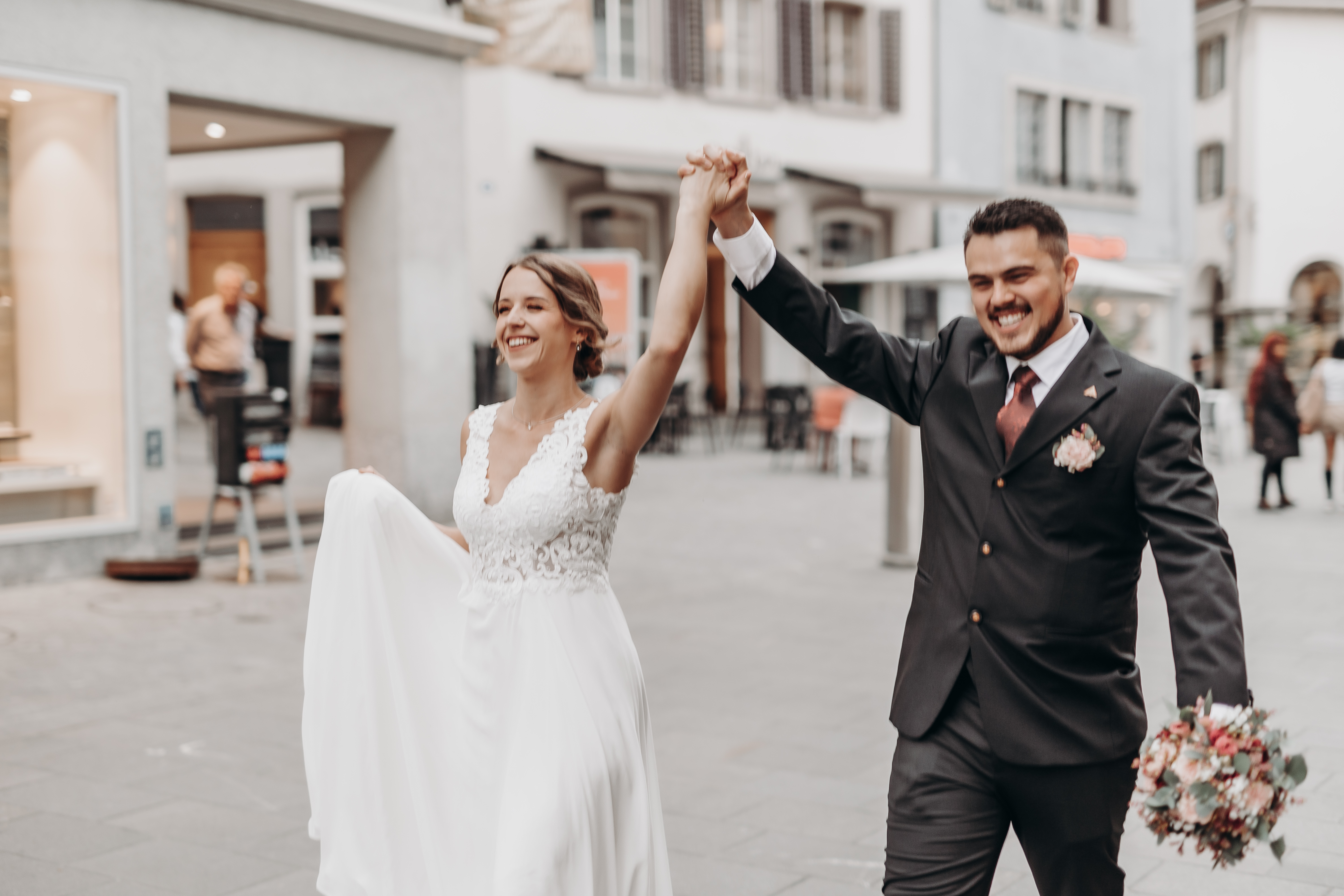 Eine fröhliche Braut und ein Bräutigam gehen Hand in Hand durch eine Badener Stadtstraße. Die Braut trägt ein weißes Spitzenkleid und hält ihr Kleid mit einer Hand, während der Bräutigam, in einem schwarzen Anzug mit einer rosa Boutonniere gekleidet, ihre gefalteten Hände triumphierend in die Höhe streckt. Ihre Ziviltrauung wurde von der Hochzeitsfotografin wunderschön festgehalten.