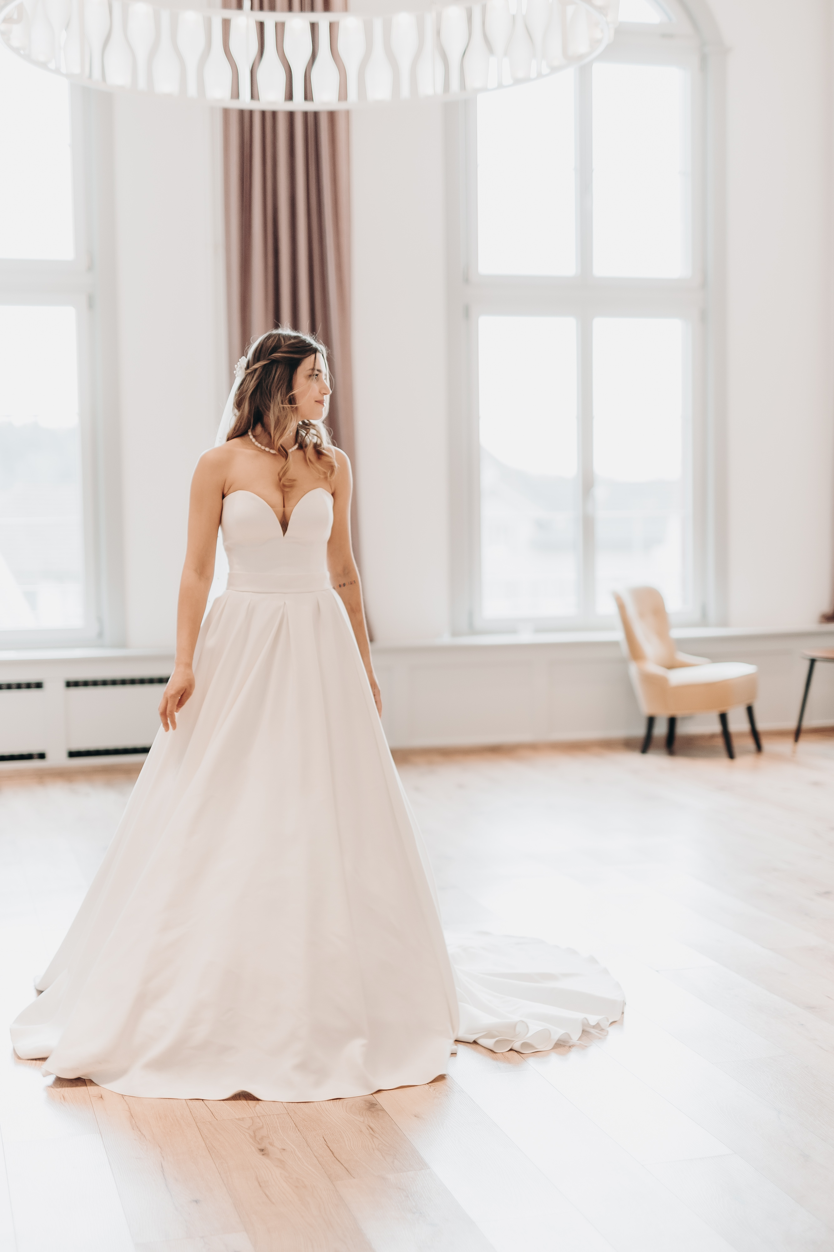 Eine Braut in einem schulterfreien, weißen Brautkleid mit langer Schleppe steht in einem hellen Raum mit großen Fenstern und hellem Holzboden. Sie blickt zur Seite, umgeben von einem beigen Stuhl, grauen Vorhängen und der natürlichen Schönheit des Aargaus, wunderschön eingefangen von Tasha Ender Photography's Hochzeitsfotografie.