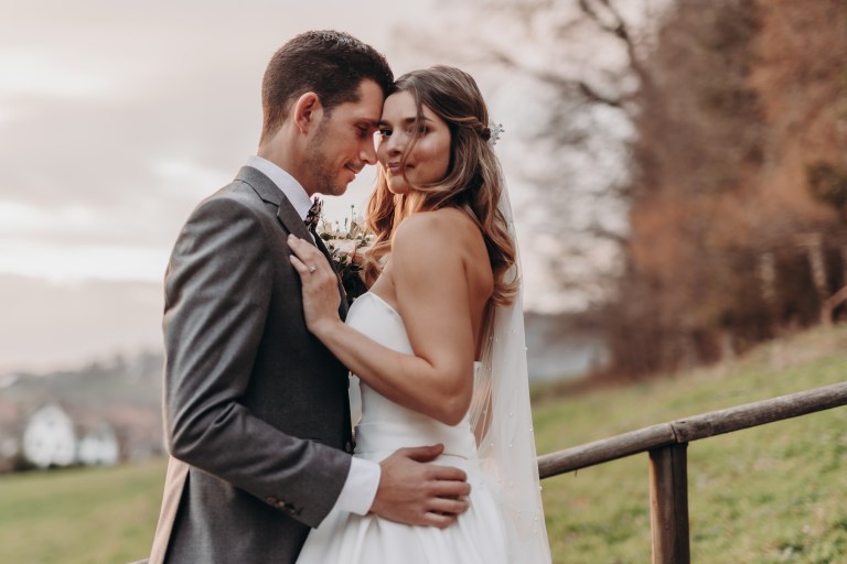 Braut und Bräutigam umarmen sich an ihrem Hochzeitstag im Freien in Baden, Aargau, aufgenommen vom Hochzeitsfotografie-Service von Tasha Ender Photography. Die Braut trägt ein weißes Kleid und einen Schleier, während der Bräutigam einen grauen Anzug trägt. Vor dem Hintergrund von üppigem Grün und einem teilweise bewölkten Himmel blicken sie sich liebevoll an und schaffen so eine mühelos schöne Szene.