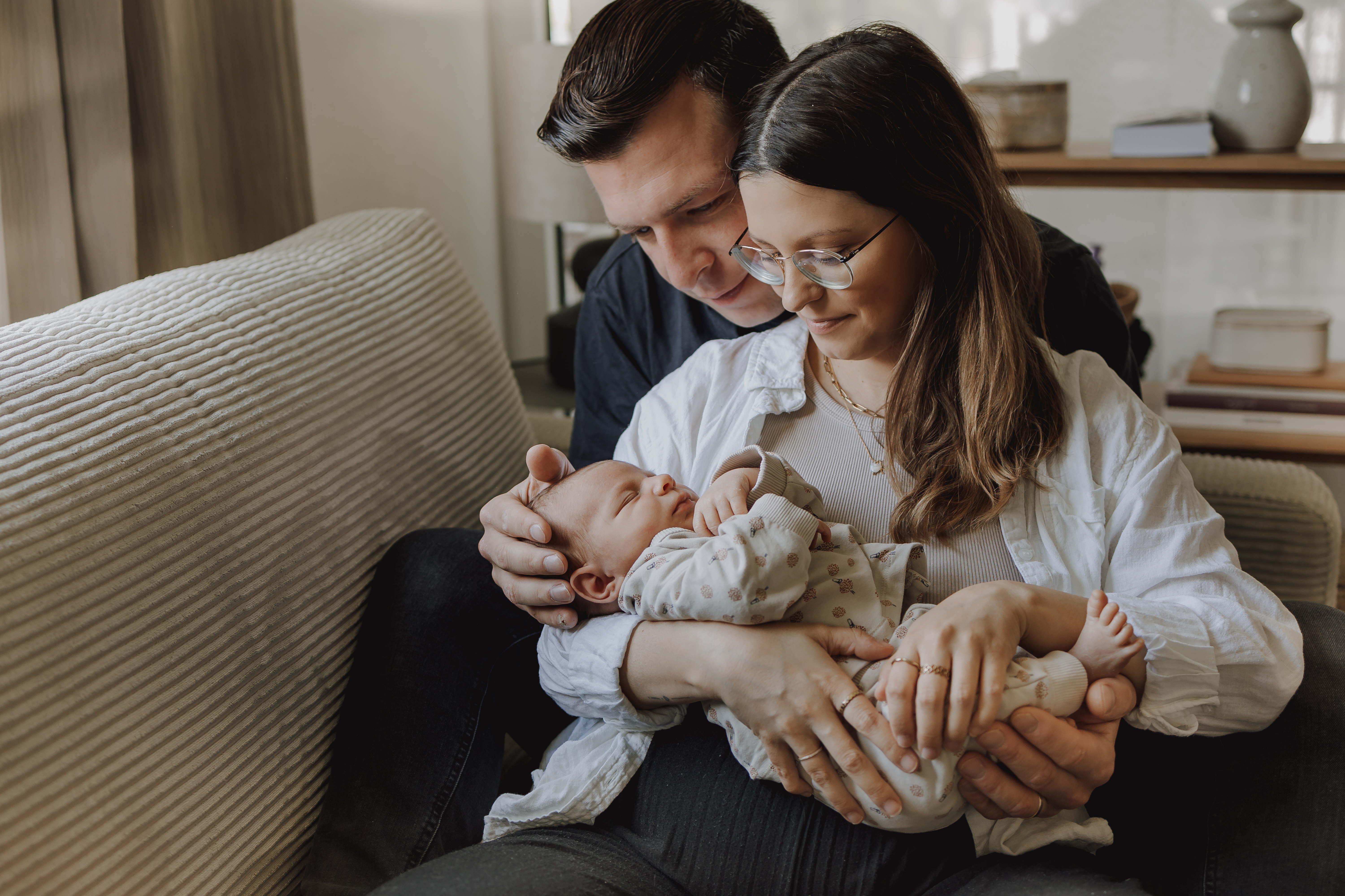 Ein Paar sitzt auf einer Couch und hält liebevoll ein schlafendes Baby im Arm. Die Frau, die eine Brille und eine weiße Bluse trägt, lächelt beim Newbornshooting bei Zuhause auf das Baby herab, während der Mann, dunkel gekleidet, sie mit zärtlichem Gesichtsausdruck von hinten umarmt.