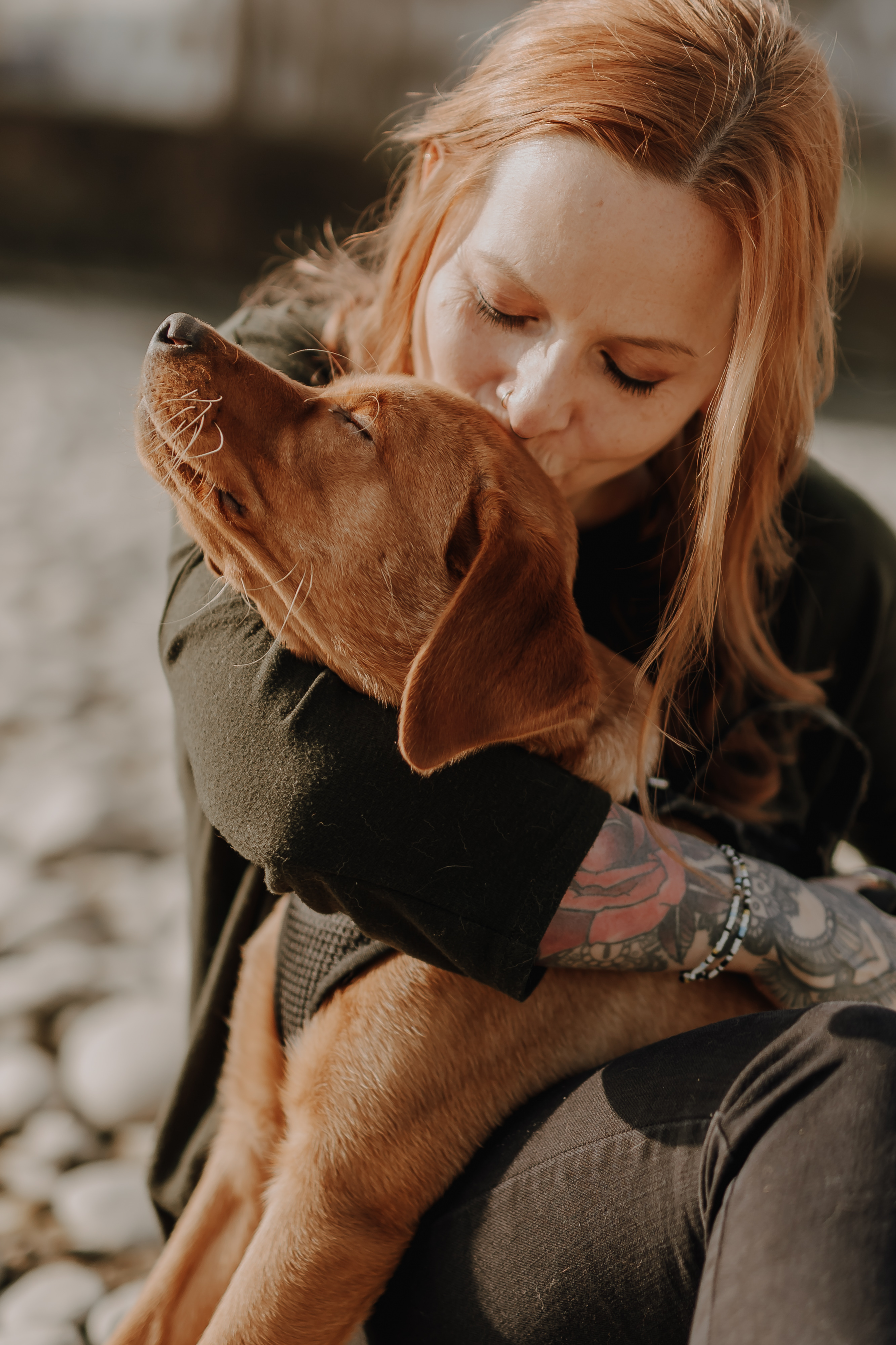 Eine Frau mit langen, rötlichen Haaren und Tattoos auf dem Arm umarmt und küsst zärtlich einen braunen Hund. Sie sind draußen an einem Kieselstrand. Aufgenommen von einem talentierten Tierfotografen wirkt der Hund zufrieden und entspannt mit geschlossenen Augen.