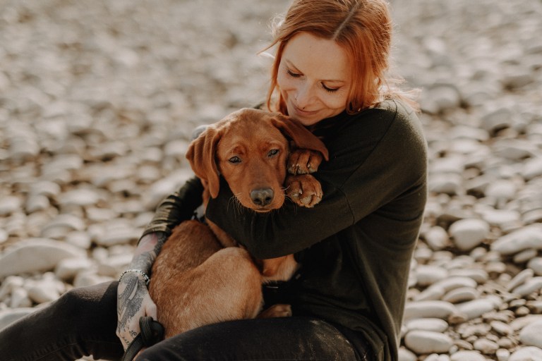 Eine Person mit langen roten Haaren und einem tätowierten Arm, möglicherweise ein talentierter Hundefotograf, sitzt an einem steinigen Strand, umarmt und lächelt einen braunen Welpen auf ihrem Schoß an. Der Welpe wirkt entspannt und zufrieden, während er sich an die Person lehnt. Der Hintergrund ist mit glatten, sonnenbeschienenen Kieselsteinen gefüllt.