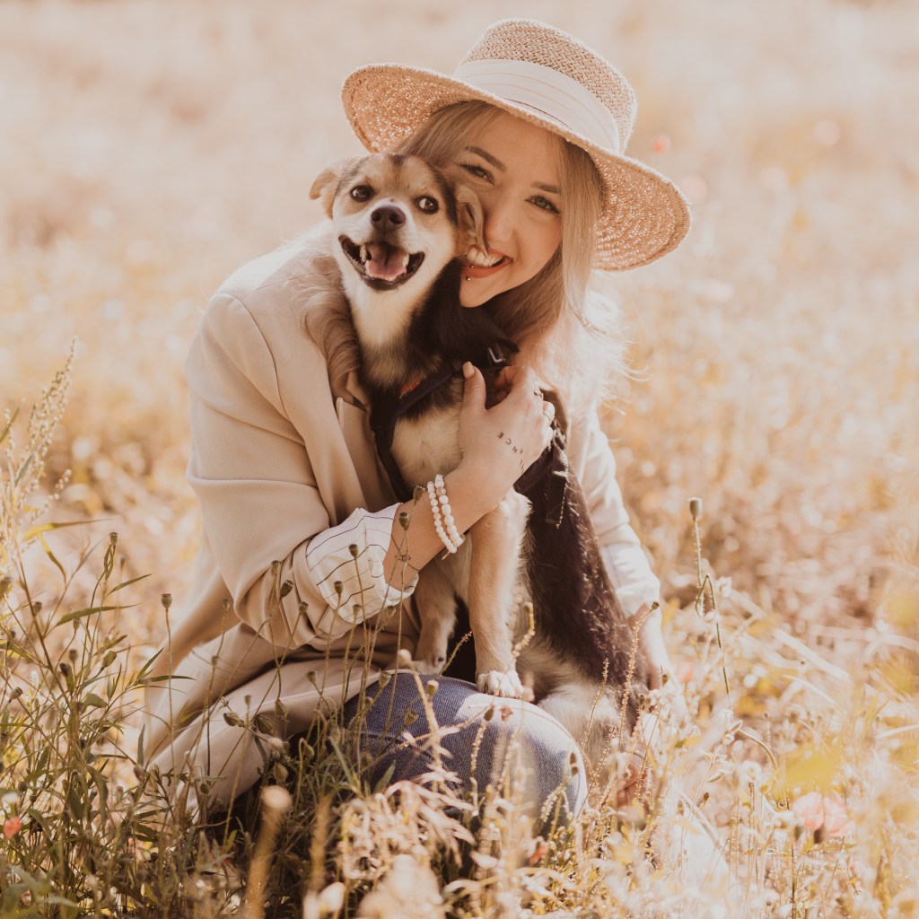 Eine lächelnde Frau mit Strohhut umarmt einen glücklichen Hund auf einer sonnigen, goldenen Wiese und fängt die Essenz der Hundefotografie ein. Im Gras sitzend, mit einer hellen Jacke und Perlenarmbändern, spiegeln beide einen freudigen Moment im Freien wider, den jeder Tierfotograf schätzen würde.