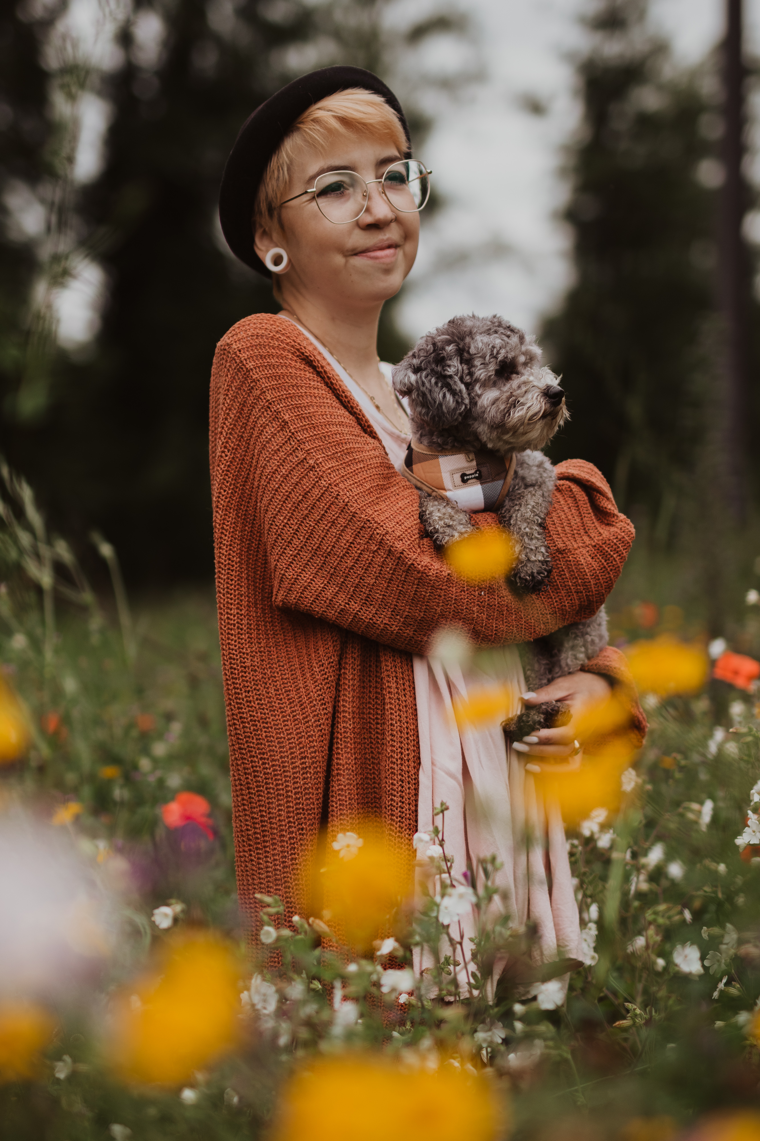 Eine Person mit Brille, schwarzer Baskenmütze und orangefarbener Strickjacke hält einen kleinen, lockigen Hund. Sie stehen auf einem Feld mit bunten Wildblumen und Grün in Baden und wirken zufrieden und friedlich. Der Hintergrund ist unscharf und hebt die Person und ihren Hund hervor, als wäre es ein professionelles Fotoshooting.