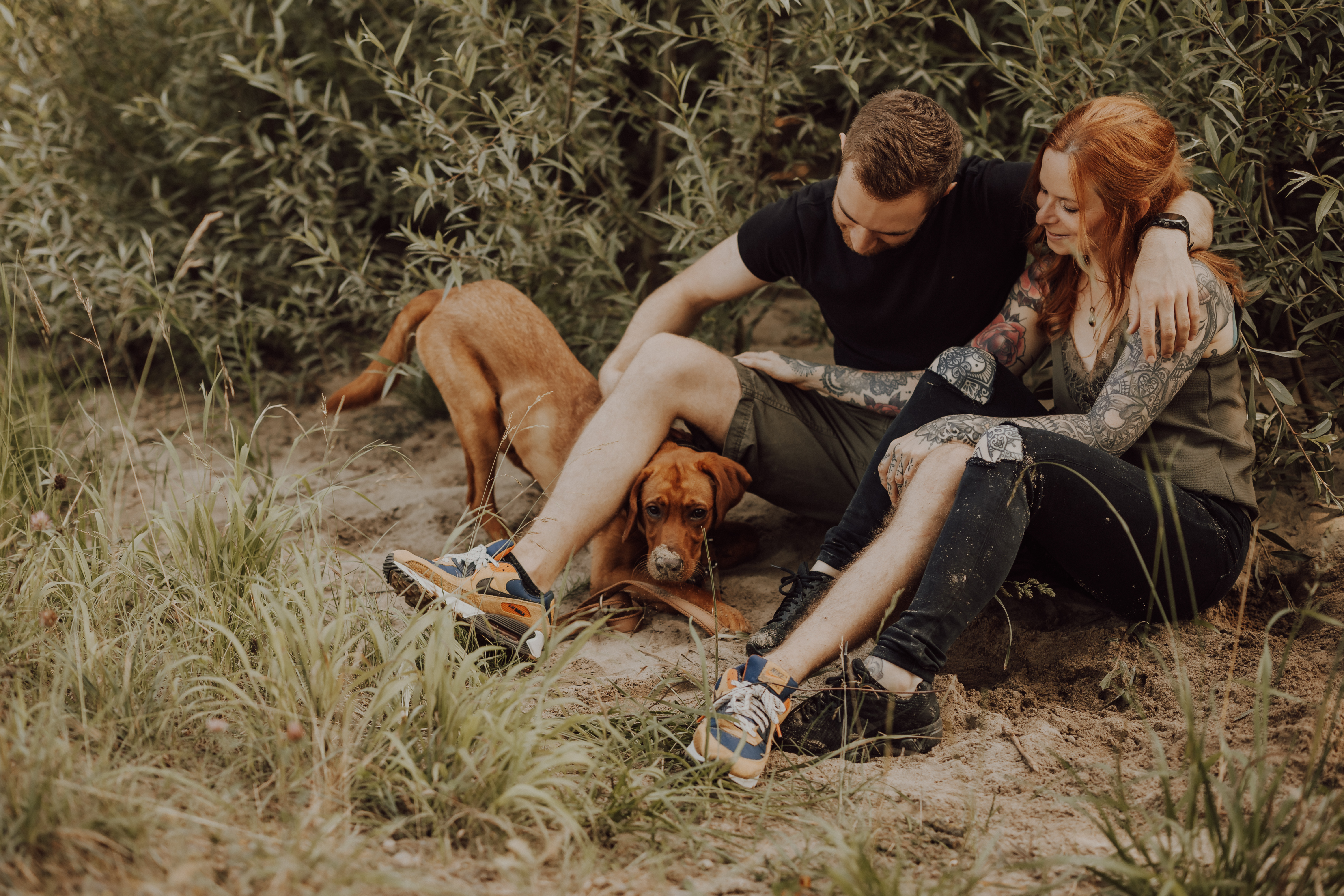 Ein Paar sitzt eng beieinander auf dem Boden im Freien, im Schatten üppiger grüner Blätter. Beide lächeln herzlich. Zwischen ihnen streckt sich ein brauner Hund glücklich im Gras aus und trägt zur heiteren und fröhlichen Atmosphäre dieses badischen Moments bei, der von einem begnadeten Tierfotografen festgehalten wurde.