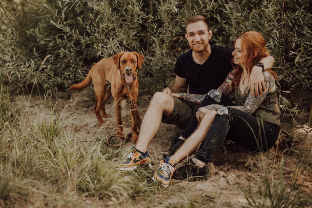 Ein lächelndes Paar sitzt draußen auf dem Boden. Der Mann trägt ein schwarzes T-Shirt und Shorts und die tätowierte Frau ein grünes Oberteil. Ihr brauner Hund, wunderschön fotografiert von einem talentierten Hundefotografen, steht an der Leine neben ihnen. Der Hintergrund ist mit üppigem grünem Laub bedeckt.