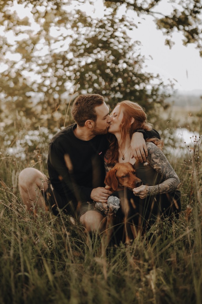 Ein Paar sitzt auf einer Wiese mit Bäumen im Hintergrund und küsst sich. Der Mann hat seinen Arm um die Frau gelegt, die lange, rote Haare und Tattoos auf den Armen hat. Sie halten einen braunen Hund. Die Szene wurde von einem talentierten Tierfotografen aufgenommen und ist in warmes, natürliches Licht getaucht, was eine heitere Atmosphäre schafft.