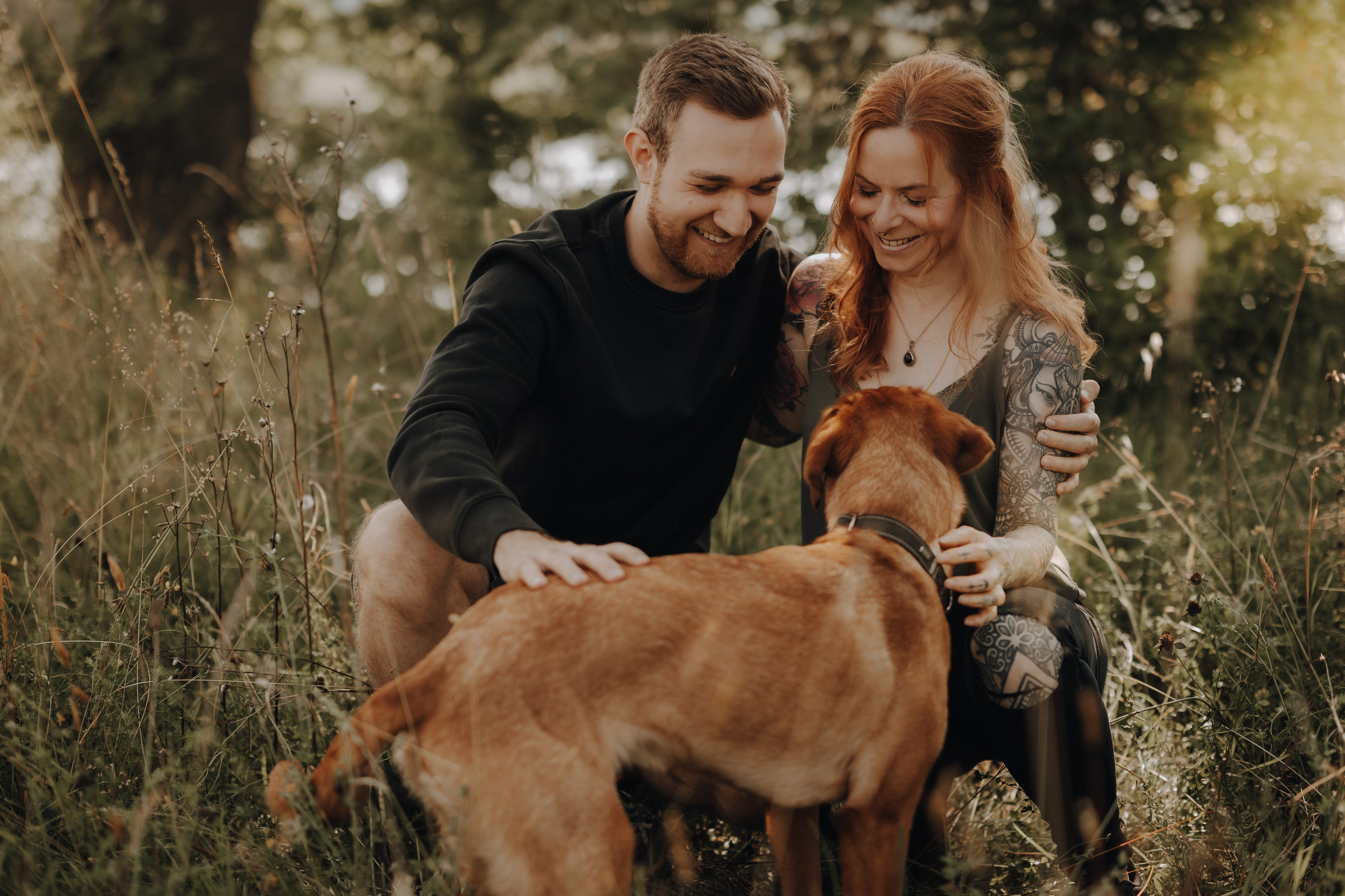 Ein Mann und eine Frau, beide lächelnd, knien auf einer Wiese und streicheln einen mittelgroßen braunen Hund. Die Frau hat Tattoos auf den Armen und sie scheinen einen schönen, sonnigen Tag im Freien mit Bäumen im Hintergrund zu genießen – ein perfekter Moment, eingefangen von Hundefotografie.