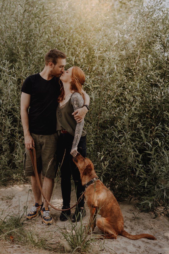 Ein Paar umarmt und küsst sich, während es auf einem Sandweg steht, der von üppigem Grün umgeben ist. Die Frau mit Tätowierungen auf den Armen hält eine Hundeleine. Ein Hund sitzt zu ihren Füßen und schaut liebevoll zu ihnen auf. Das Sonnenlicht fällt durch die Bäume darüber und wurde von der auf Hundefotografie spezialisierten Fotografin wunderschön eingefangen.