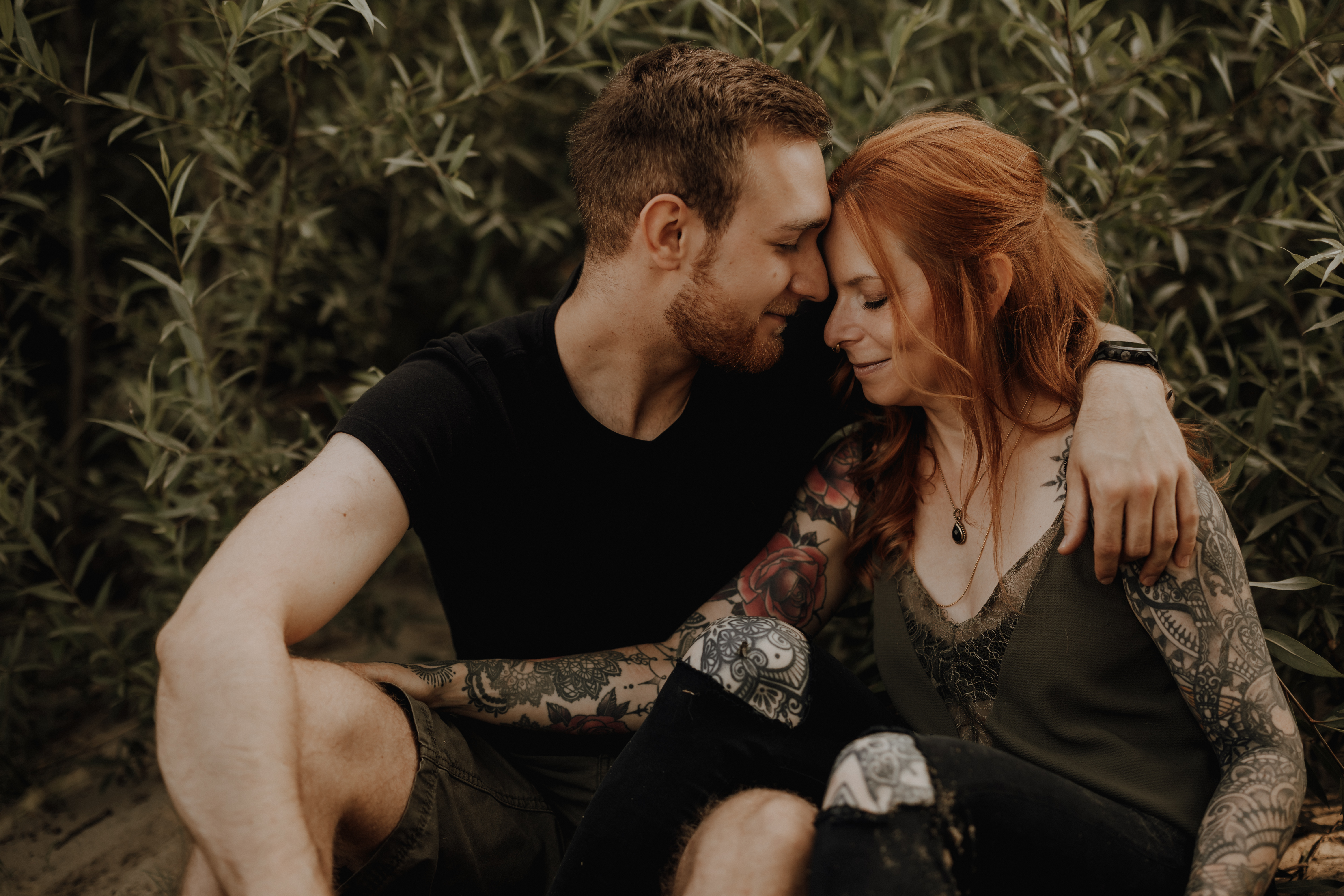 Ein Paar sitzt eng beieinander im Freien, umgeben von Grün. Sie berühren zärtlich ihre Stirnen, der Arm des Mannes liegt um die Schultern der Frau. Aufgenommen während eines Fotoshootings mit einer talentierten Fotografin, wirken beide entspannt und lächelnd, während die Frau aufwendige Tattoos auf ihren Armen zeigt.