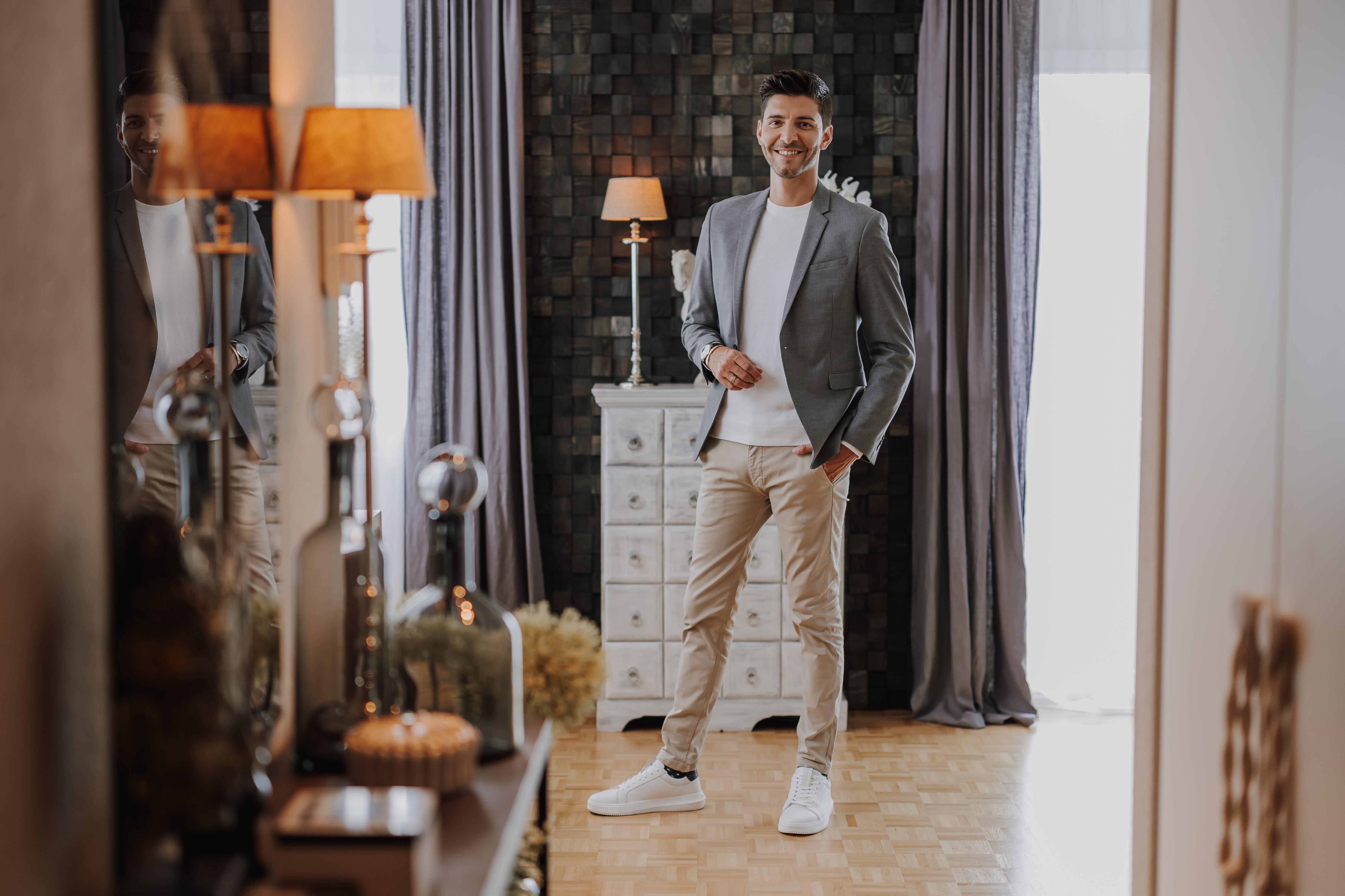 Ein Mann steht in einem stilvollen, modernen Raum mit Holzboden und lächelt. Er trägt einen grauen Blazer, einen weißen Pullover, beige Hosen und weiße Turnschuhe. Während dieser Fotoshooting-Session sind im Hintergrund ein hoher Schrank mit Schubladen und dekorativen Lampen sowie bodenlange Vorhänge zu sehen.