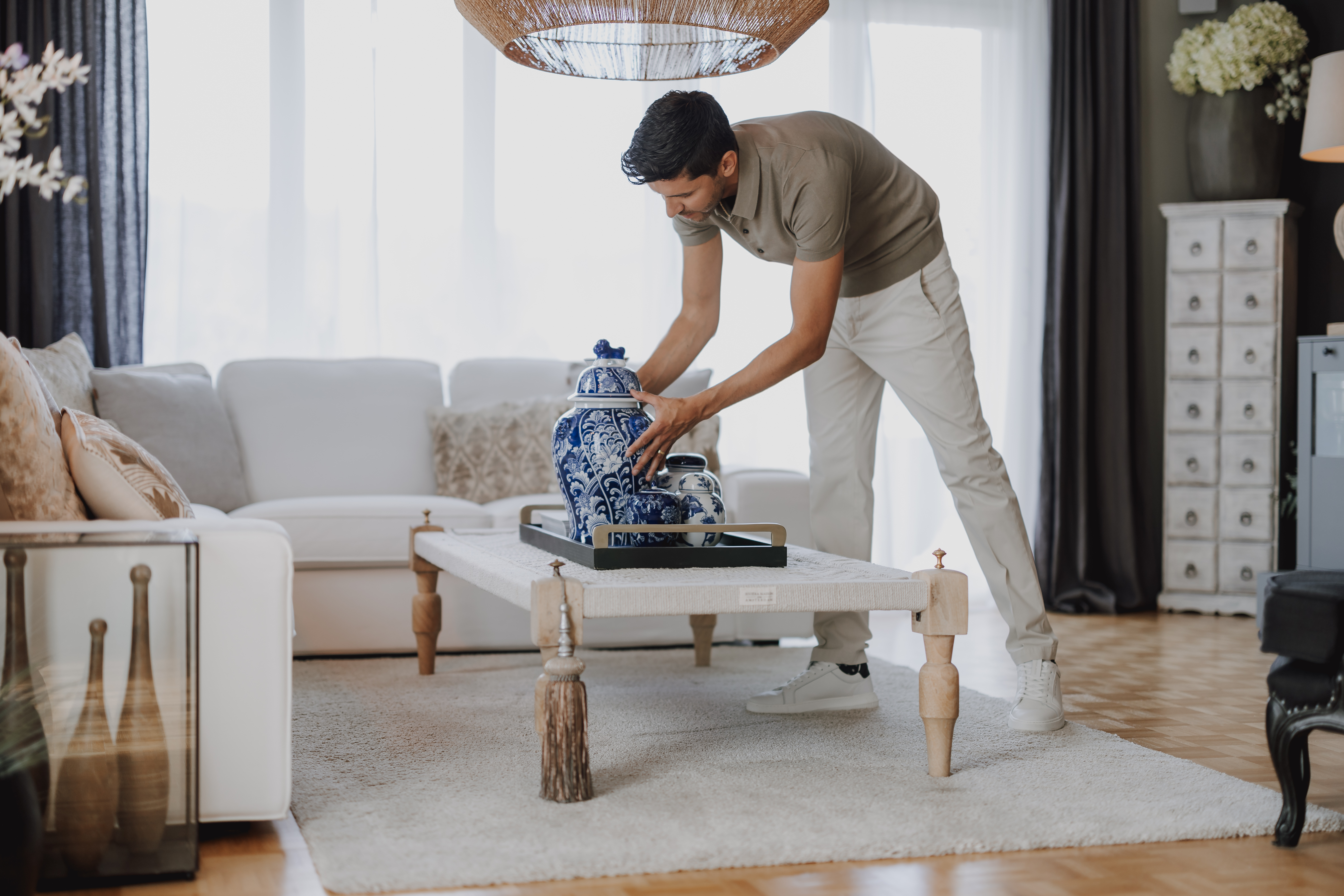 Ein Mann arrangiert blaue und weiße Keramikvasen auf einem Couchtisch in einem stilvoll eingerichteten Wohnzimmer. Der Raum verfügt über ein weißes Sofa, einen hellgrauen Teppich, moderne Möbel und Holzböden. Große Fenster füllen den Raum mit natürlichem Licht, perfekt für ein Fotoshooting oder zum Erstellen von Inhalten für Ihre Website.