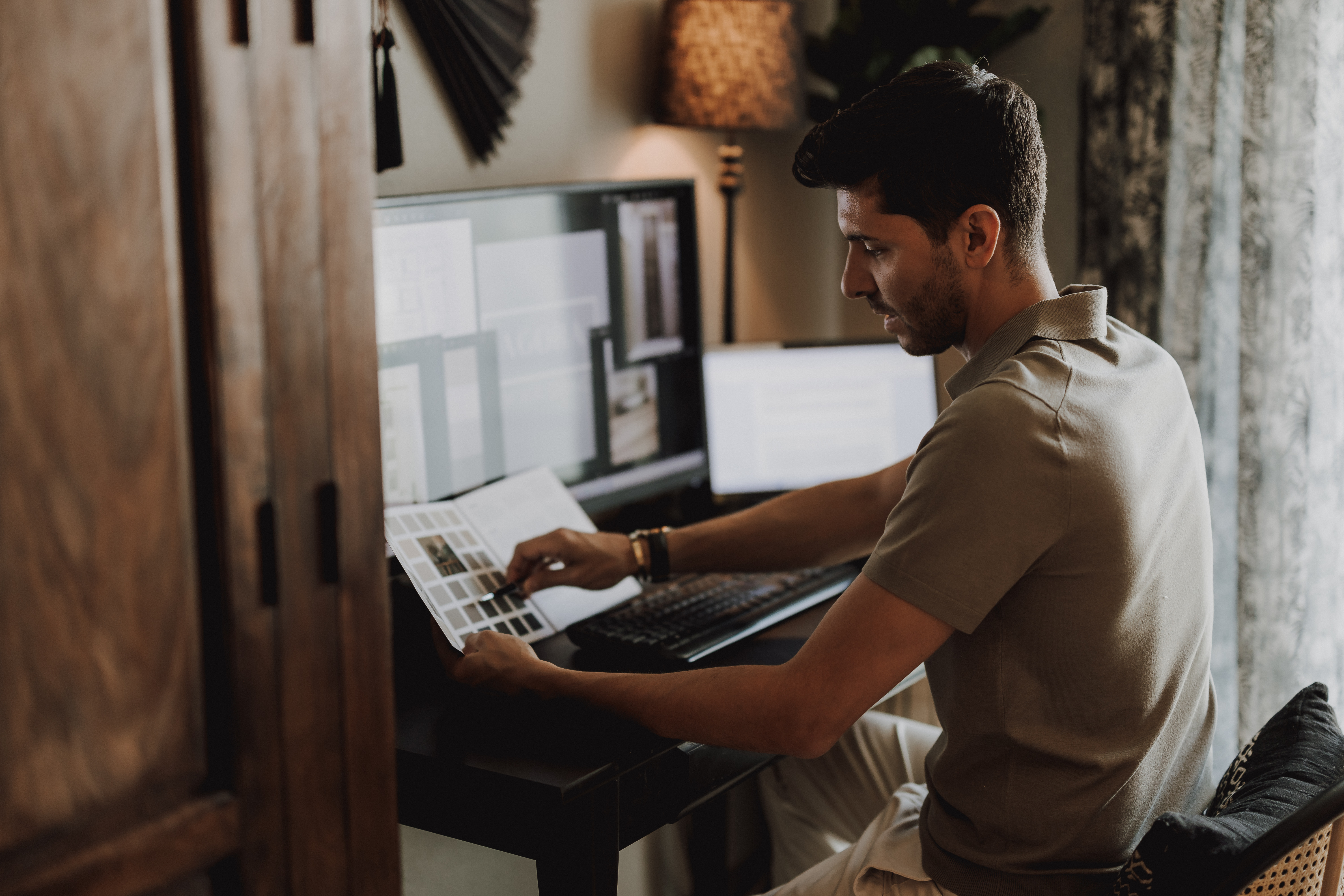Ein Mann sitzt an einem Schreibtisch in einem Home-Office und ist auf seine Arbeit konzentriert. Er betrachtet ein Farbmuster, während auf zwei Computermonitoren verschiedene Fenster angezeigt werden. Während er gerade ein Fotoshooting für seine kommende Webseite macht, ist die Umgebung durch eine Lampe auf dem Schreibtisch und teilweise zugezogene Vorhänge vor einem Fenster warm beleuchtet.