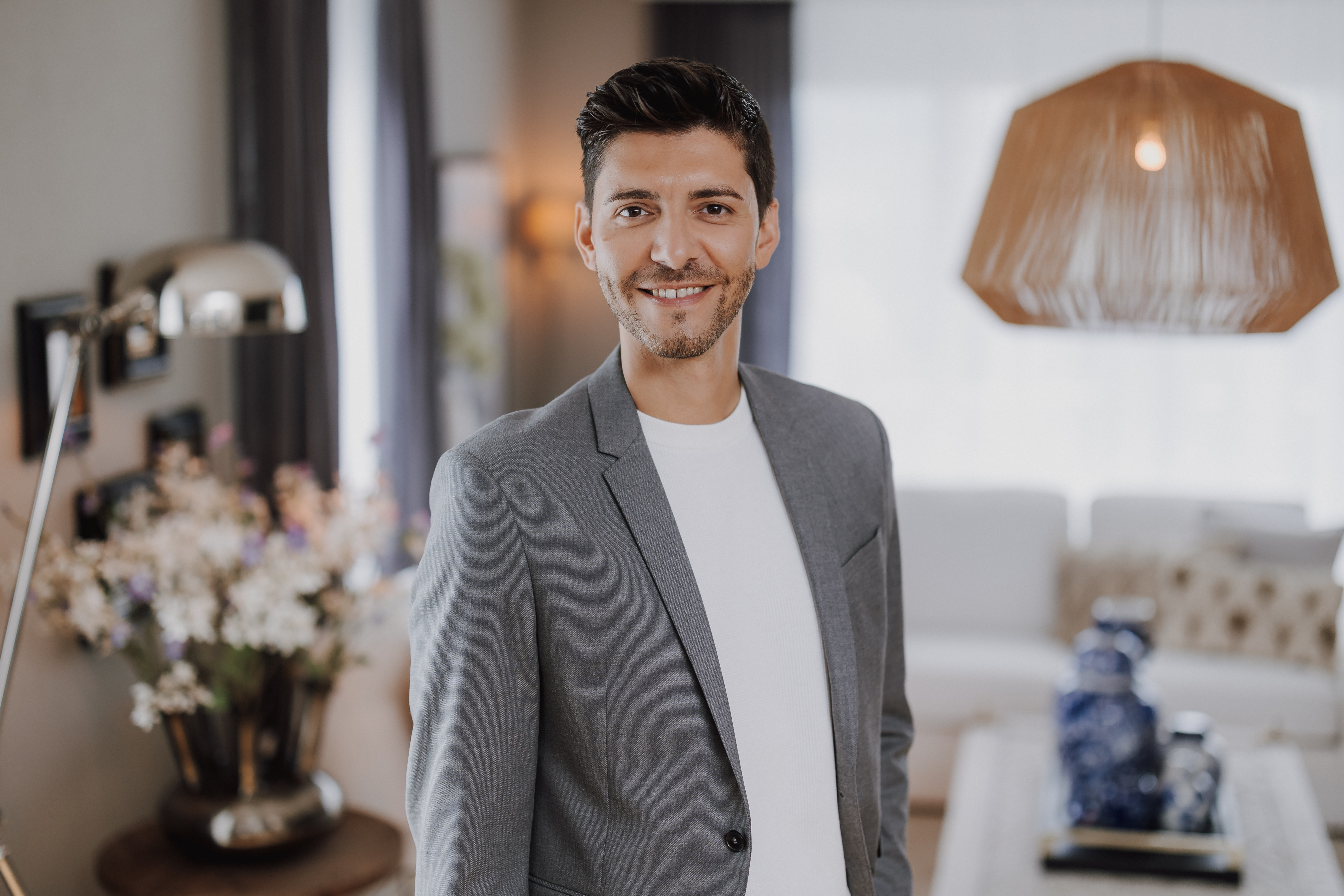 Ein Mann mit kurzen dunklen Haaren und gestutztem Bart lächelt. Er trägt einen grauen Blazer über einem weißen Hemd, perfekt für Fotos für Webseite. Der Hintergrund zeigt einen modernen, gut beleuchteten Raum mit einer Lampe, Blumen in einer Vase und einer dekorativen Leuchte, die jeder Fotograf für ein Reportage-Shooting lieben würde.
