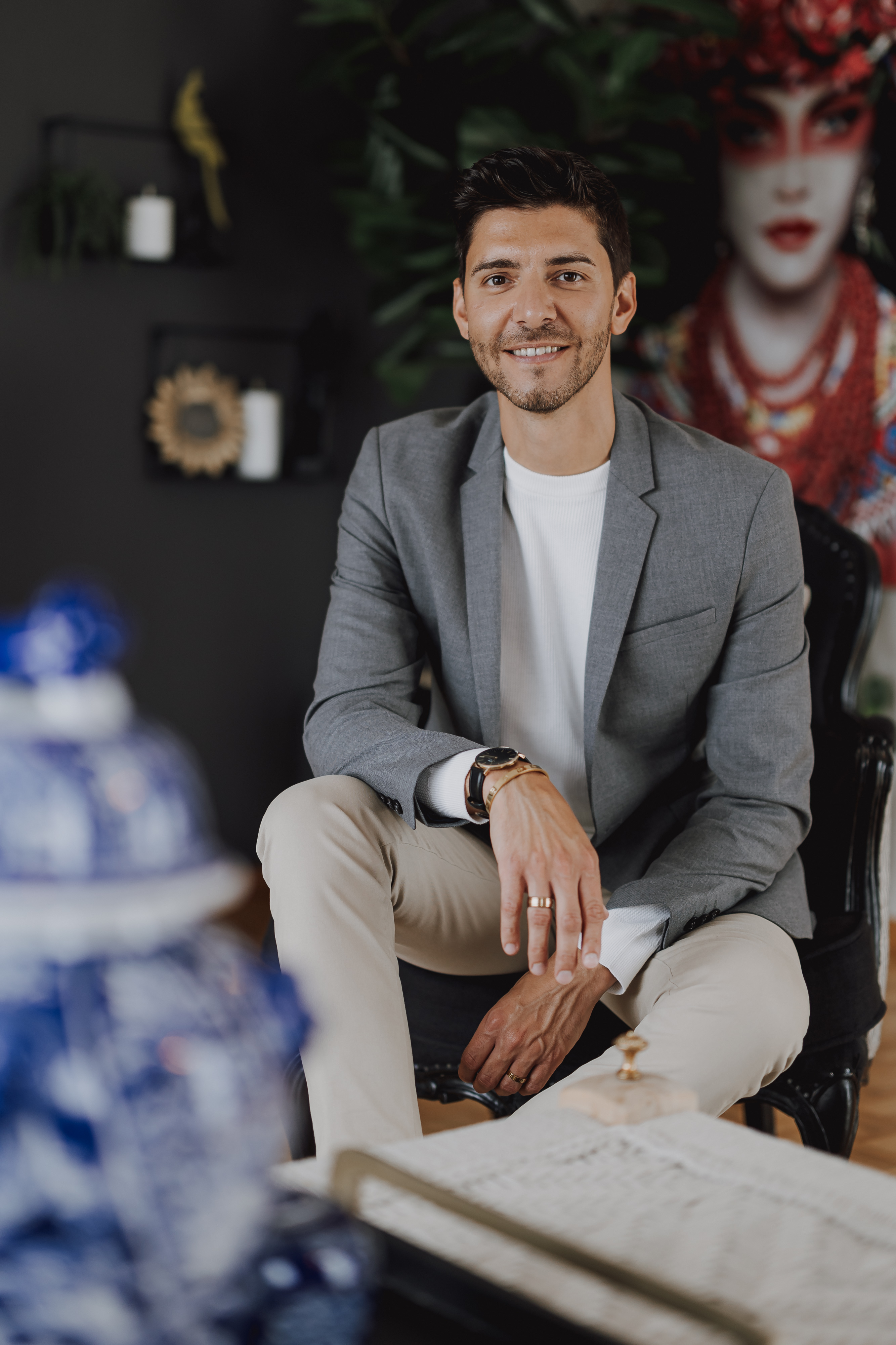 Ein Mann mit dunklem Haar und Bart, der einen grauen Blazer und cremefarbene Hosen trägt, sitzt mit entspanntem, freundlichem Gesichtsausdruck auf einem schwarzen Stuhl. Im Hintergrund ist ein großes, farbenfrohes Gemälde mit dem Gesicht einer Frau in traditioneller Kleidung und einigen Dekorationsgegenständen auf einem Regal zu sehen, wodurch ein fast reportagehafter Inhalt entsteht.