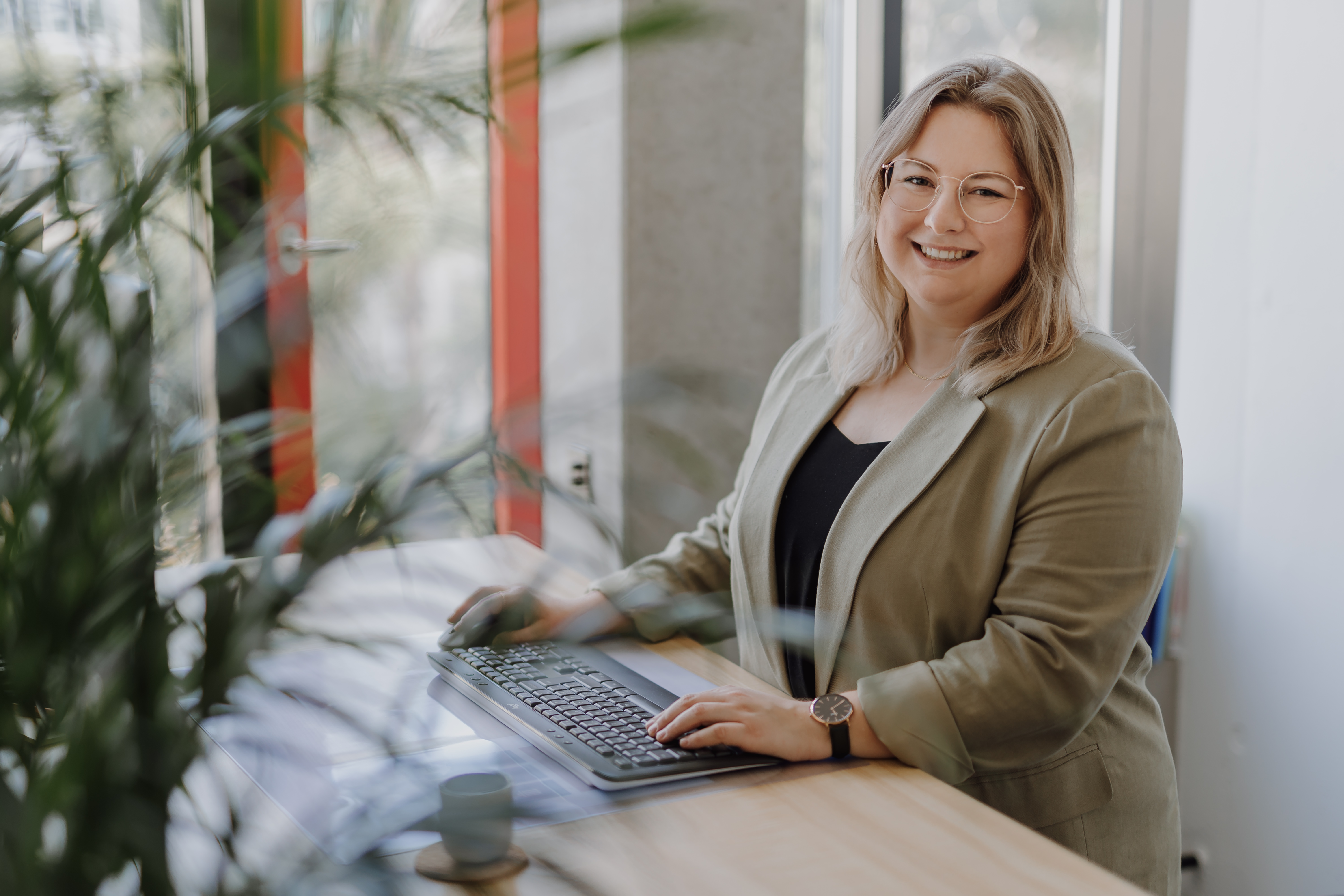 Eine Person mit schulterlangem blondem Haar und Brille sitzt lächelnd an einem Schreibtisch mit Tastatur und ist bereit für ihr CV-Fotoshooting. Sie trägt einen hellen Blazer und ein dunkles Oberteil. Im Hintergrund sind verschwommenes Grün und Fenster zu sehen, die natürliches Licht hereinlassen.