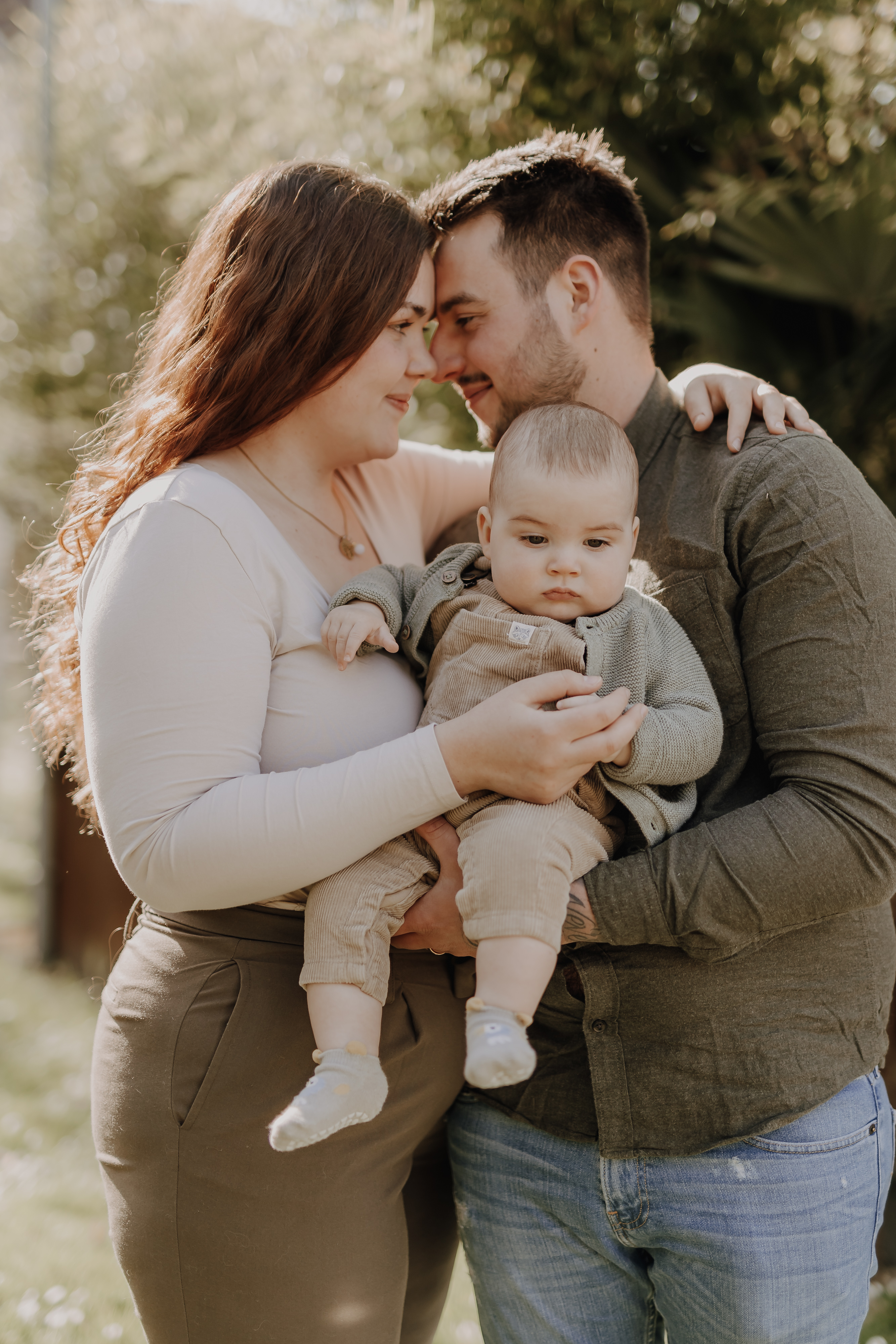 Ein Paar steht im Freien eng beieinander, umarmt sich und hält während eines herzerwärmenden Babyshootings ein Baby zwischen sich. Die Frau mit dem langen, welligen rotbraunen Haar trägt ein helles Oberteil und olivfarbene Hosen, während der Mann in einem grünen Hemd und Jeans ihr beige gekleidetes Baby liebevoll im Arm hält.