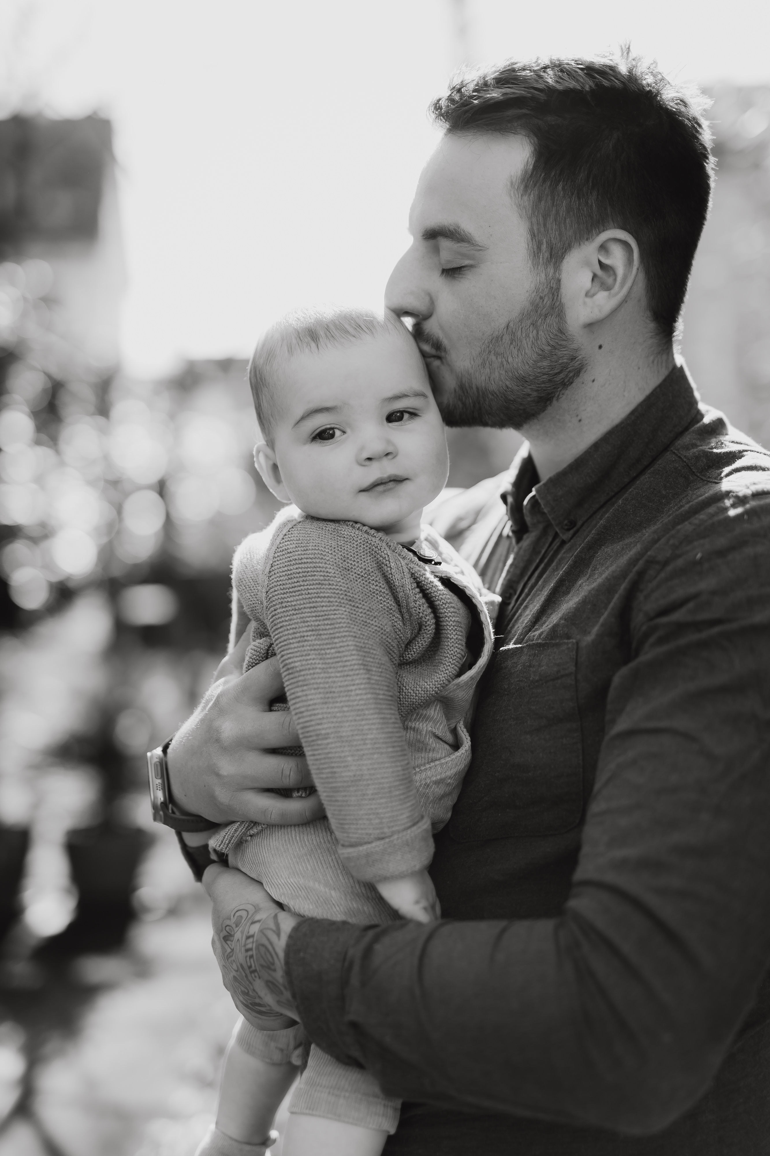 Während eines Familienshootings, das von einer talentierten Fotografin arrangiert wurde, küsst ein Mann mit Bart den Kopf eines Babys, das er in seinen Armen hält. Der Mann trägt ein langärmeliges Hemd und hat Tattoos auf der Hand. Das Baby, das einen Strickpullover trägt, blickt vor der leicht verschwommenen Außenkulisse direkt in die Kamera.