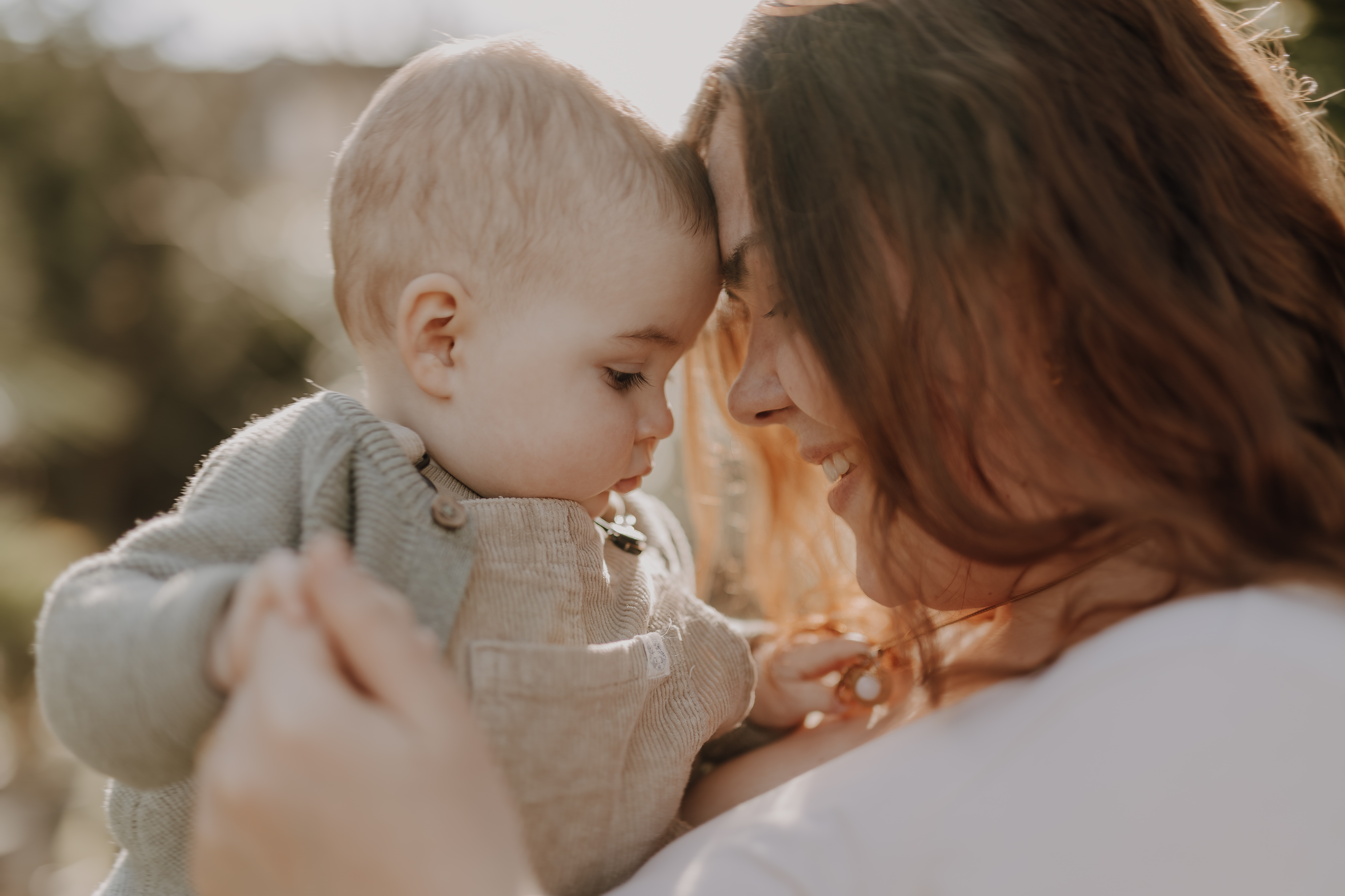 Eine Frau mit langen braunen Haaren hält ein Baby sanft an sich gedrückt, beide lächeln und berühren ihre Stirnen. Das Baby trägt einen hellgrünen Pullover über einem grauen Hemd, während die Frau ein weißes Oberteil trägt. Sie sind während eines intimen Fotoshootings draußen vor einem verschwommenen grünen Hintergrund und schaffen einen warmen, zärtlichen Moment.