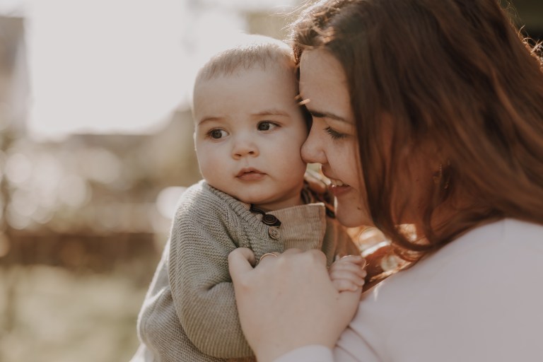 Eine Frau mit langen braunen Haaren hält und kuschelt ein Baby. Sie lächelt und schaut das Baby an, das einen grauen Pullover trägt und wegschaut. Der Hintergrund ist leicht verschwommen, was darauf schließen lässt, dass sie draußen sind und Sonnenlicht hereinströmt – ein perfekter Moment für Familienfotos oder eine süße Babyshooting-Session.