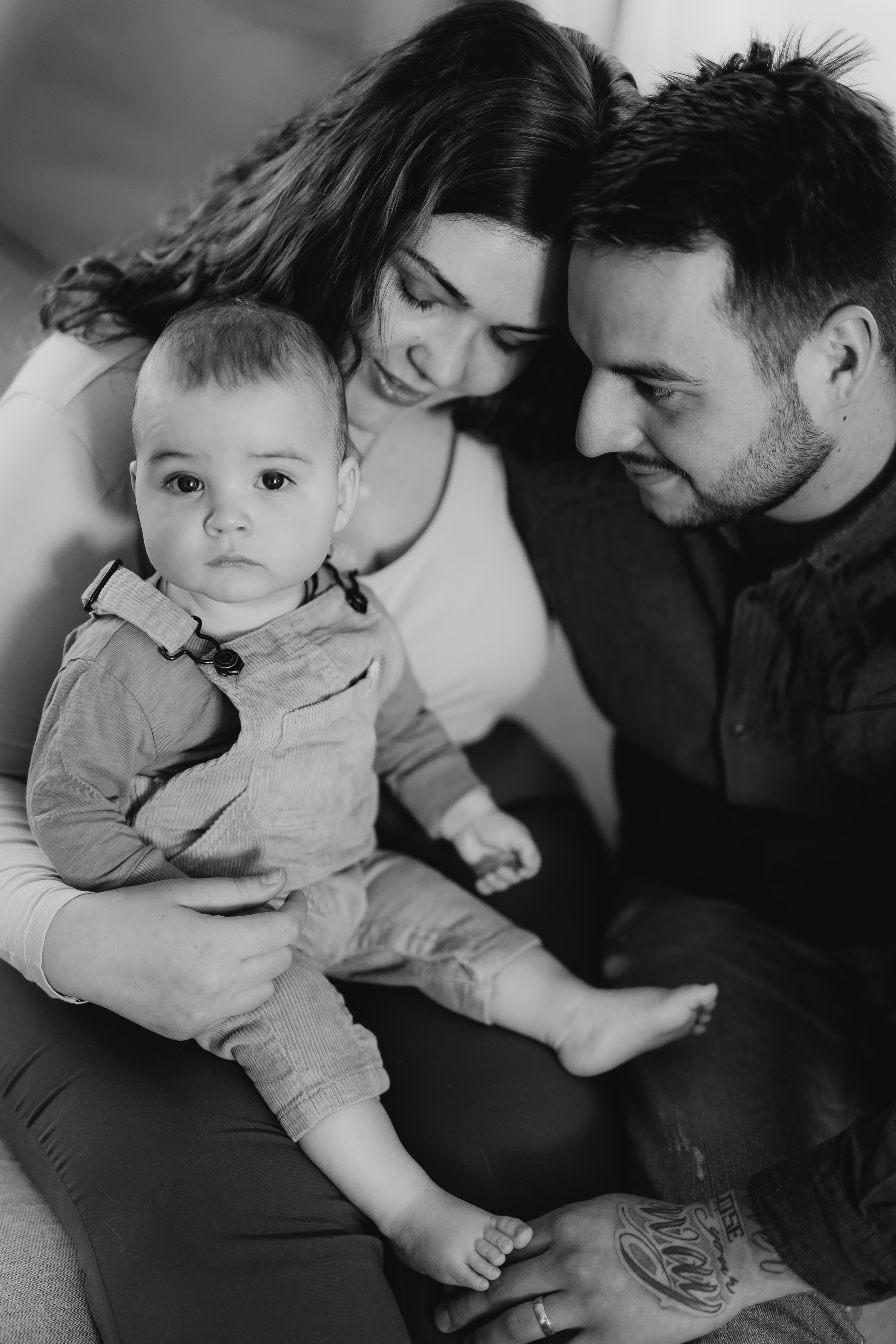Ein Schwarz-Weiß-Foto eines Paares, das eng beieinander sitzt und sein Baby hält. Das Baby, mit kurzen Haaren und Overall, sitzt auf dem Schoß der Frau, während sie, mit langen Haaren, auf das Kind herablächelt. Der Mann lächelt dem Säugling während dieses intimen Familienshootings herzlich zu.
