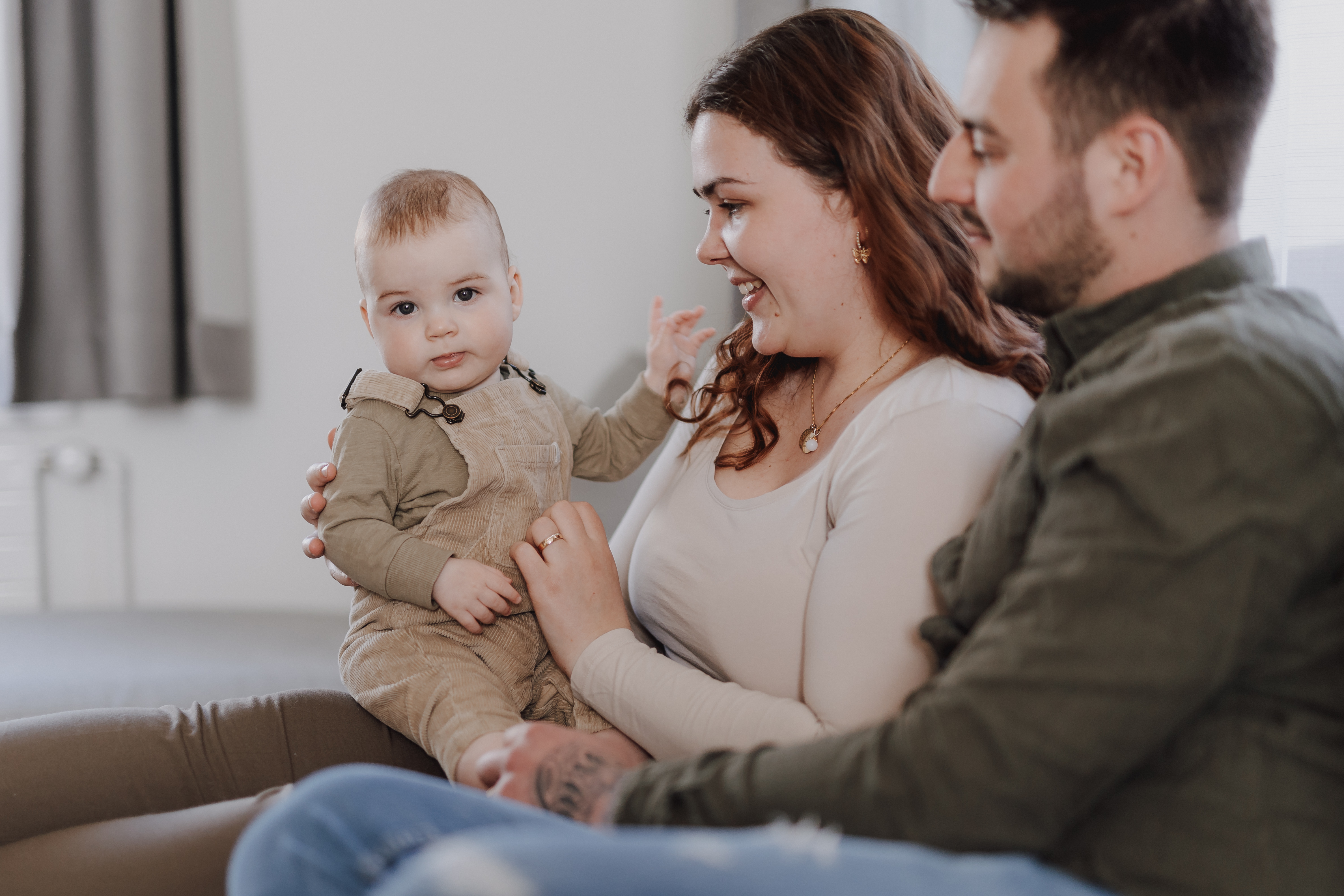 Eine glückliche Familie genießt einen gemütlichen Moment zu Hause. Bei dieser Babyshooting-Sitzung sitzt ein Baby in einem beigen Outfit auf dem Schoß einer erwachsenen Frau, die lächelt und das Baby ansieht. Ein erwachsener Mann, der ebenfalls lächelt, sitzt neben ihnen. Alle wirken zufrieden und fühlen sich wohl.