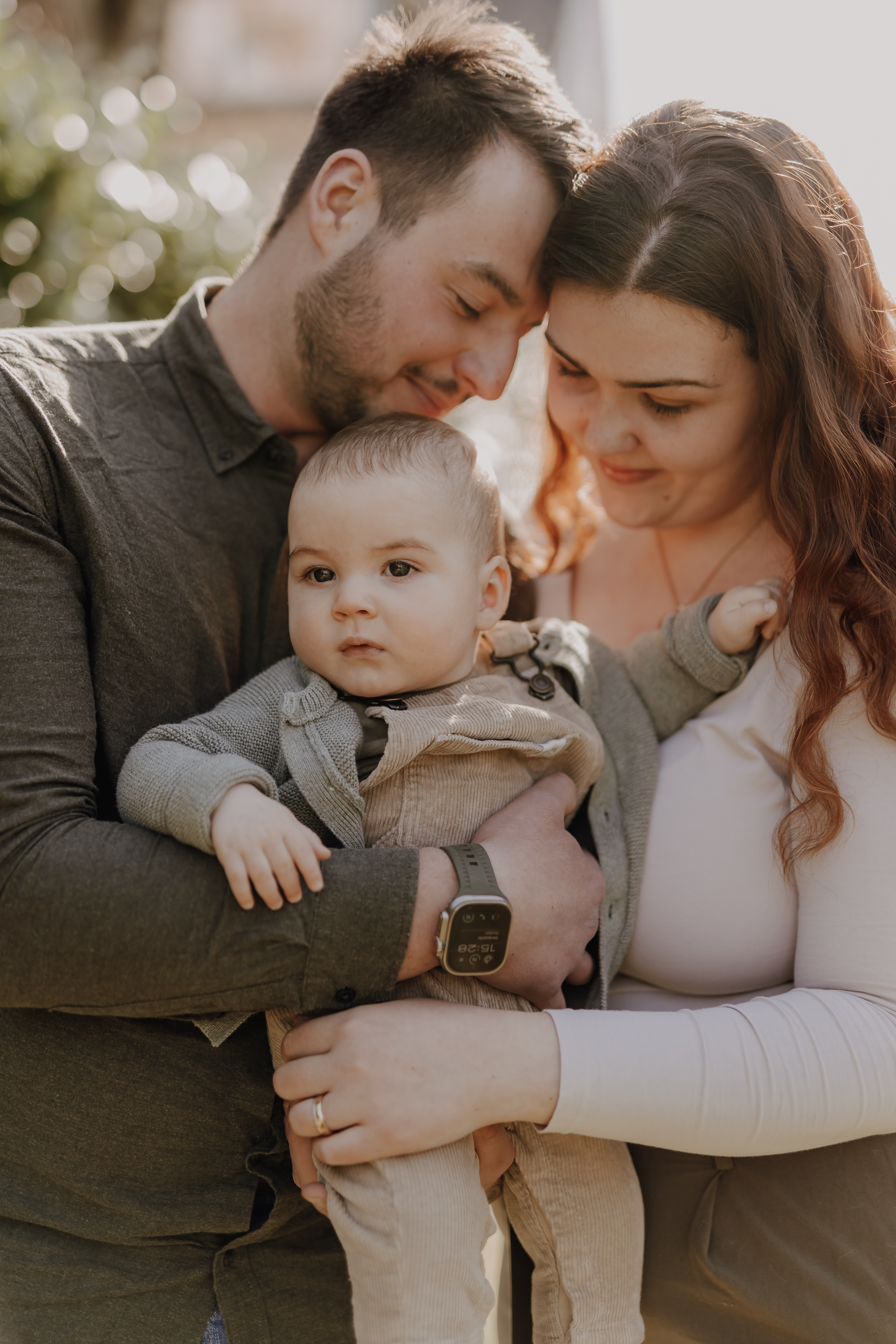 Bei einem gemütlichen Familienshooting stehen ein Mann und eine Frau eng beieinander, lächeln sich liebevoll an und halten ihr Baby im Arm. Der Mann links trägt ein dunkles Hemd, die Frau rechts ein helles Oberteil. Das Baby, hell bekleidet und mit einer grauen Strickjacke bekleidet, blickt in die Ferne.