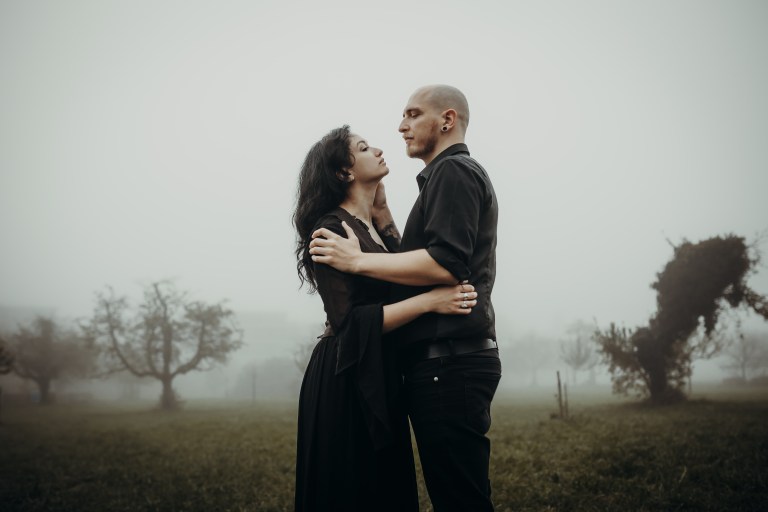 Ein Paar steht umarmend auf einem nebligen Feld. Der Mann und die Frau, in dunkle Gothic-Kleidung gekleidet, blicken sich in die Augen. Bäume mit spärlichem Laub und dichtem Nebel bilden den Hintergrund und schaffen eine mystische und intime Atmosphäre, die perfekt für Paarfotografie ist.
