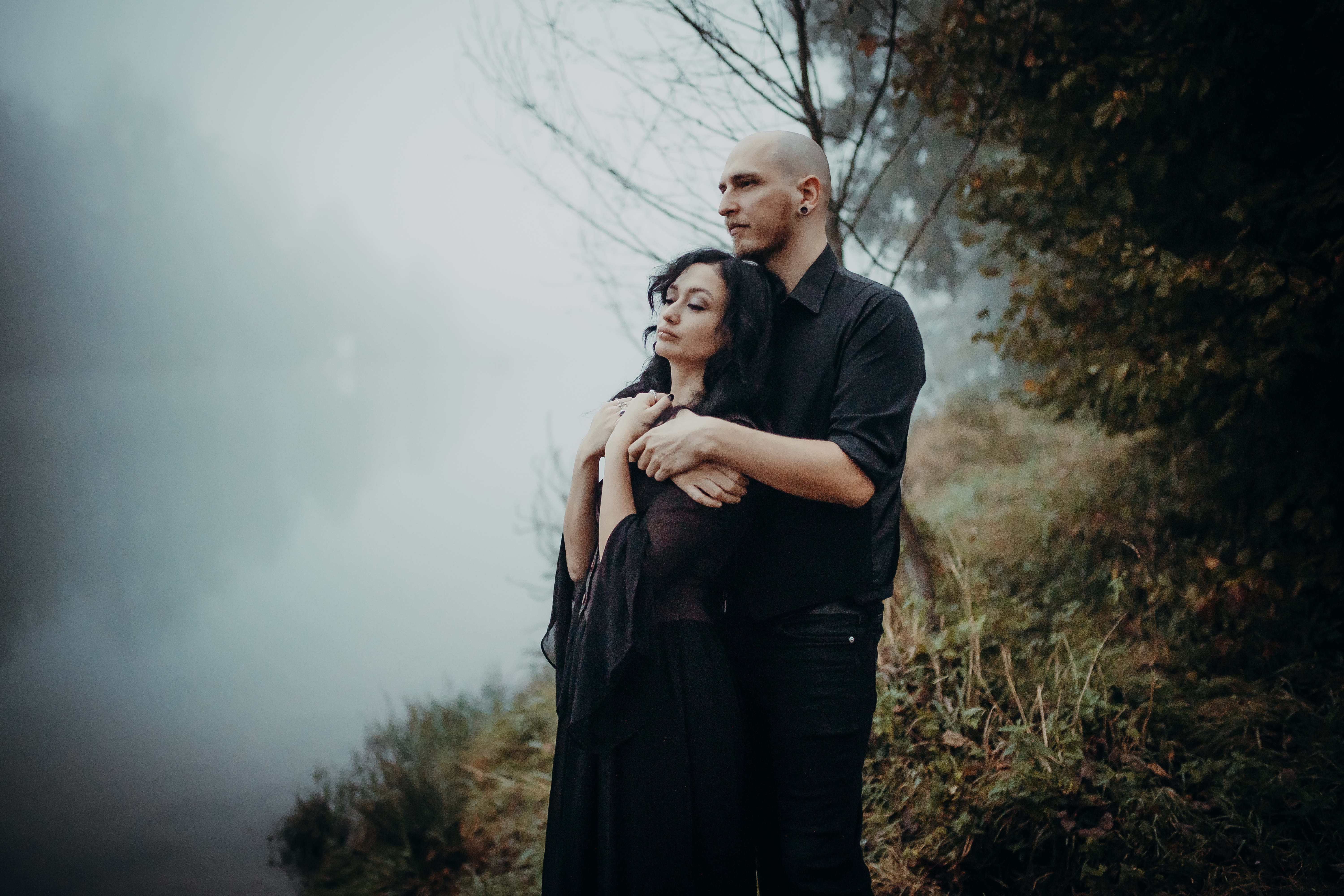 Ein Mann und eine Frau in dunkler Kleidung stehen umarmt neben einem nebligen See, umgeben von Gras und herbstlichen Bäumen. Die Frau lehnt sich mit geschlossenen Augen an die Brust des Mannes und ruft ein Gefühl von Ruhe und Intimität hervor. Nebel verdeckt die Hintergrundszene und verleiht ihrem Fotoshooting eine gotische Note.