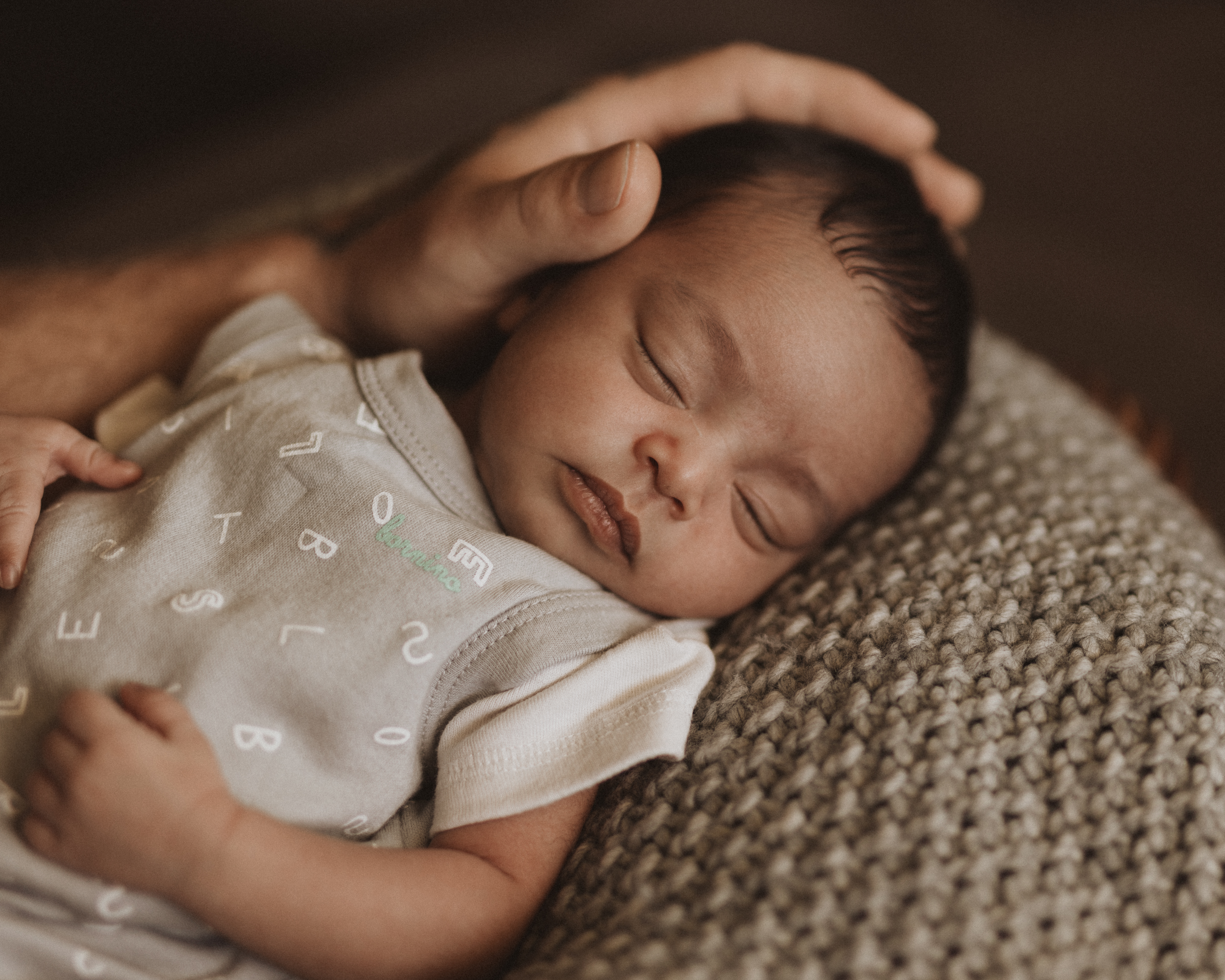Eine Nahaufnahme eines schlafenden Babys, das auf einer Strickdecke ruht. Eine erwachsene Hand wiegt sanft den Kopf des Babys. Das Baby trägt einen hellen Strampler mit aufgedruckten Buchstaben des Alphabets. Diese Babyshooting-Szene in Baden strahlt Wärme und Zärtlichkeit aus und zeigt das Können einer Neugeborenenfotografin.