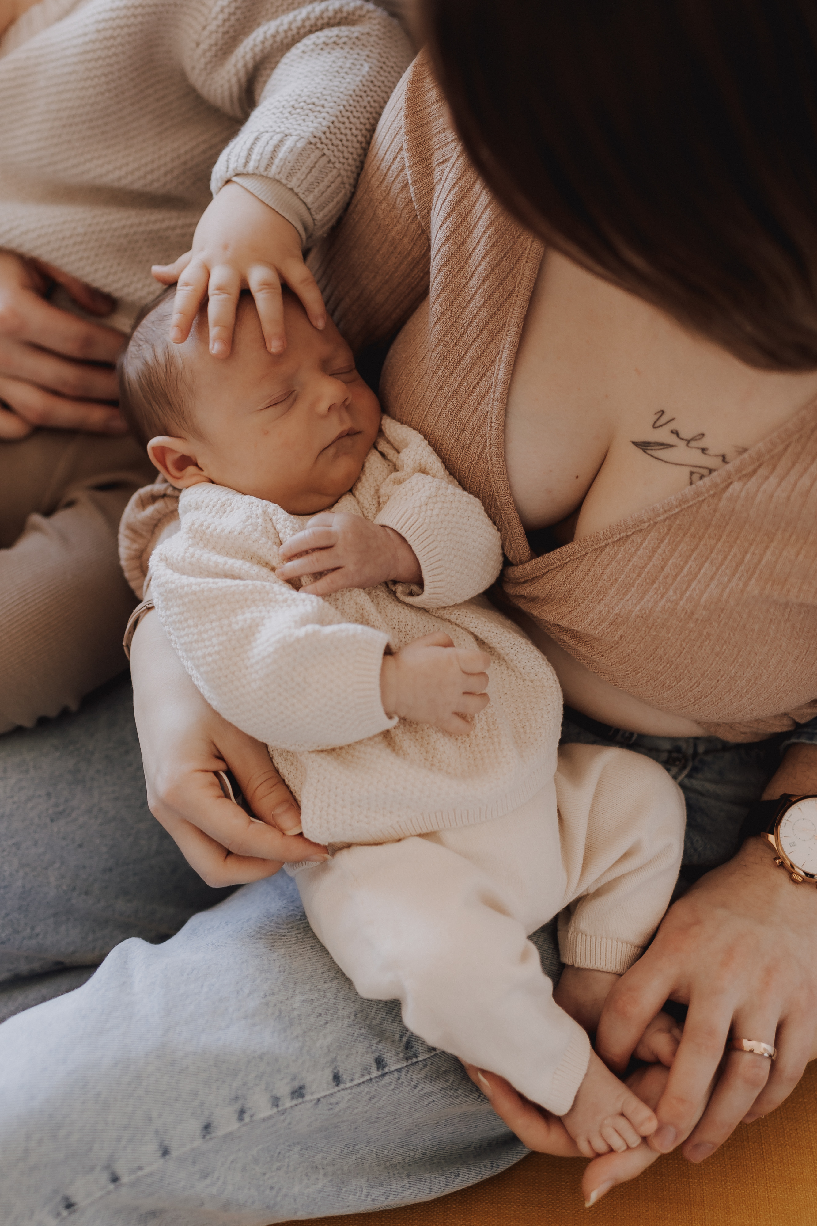 Ein schlafendes Neugeborenes, in ein cremefarbenes Outfit gekleidet, liegt in den Armen eines Erwachsenen, der einen beigen Pullover trägt. Das Baby ruht friedlich, eine Hand des Erwachsenen legt es sanft auf seinen Kopf und die andere hält es fest. So wird der perfekte Moment für ein Neugeborenenshooting festgehalten.