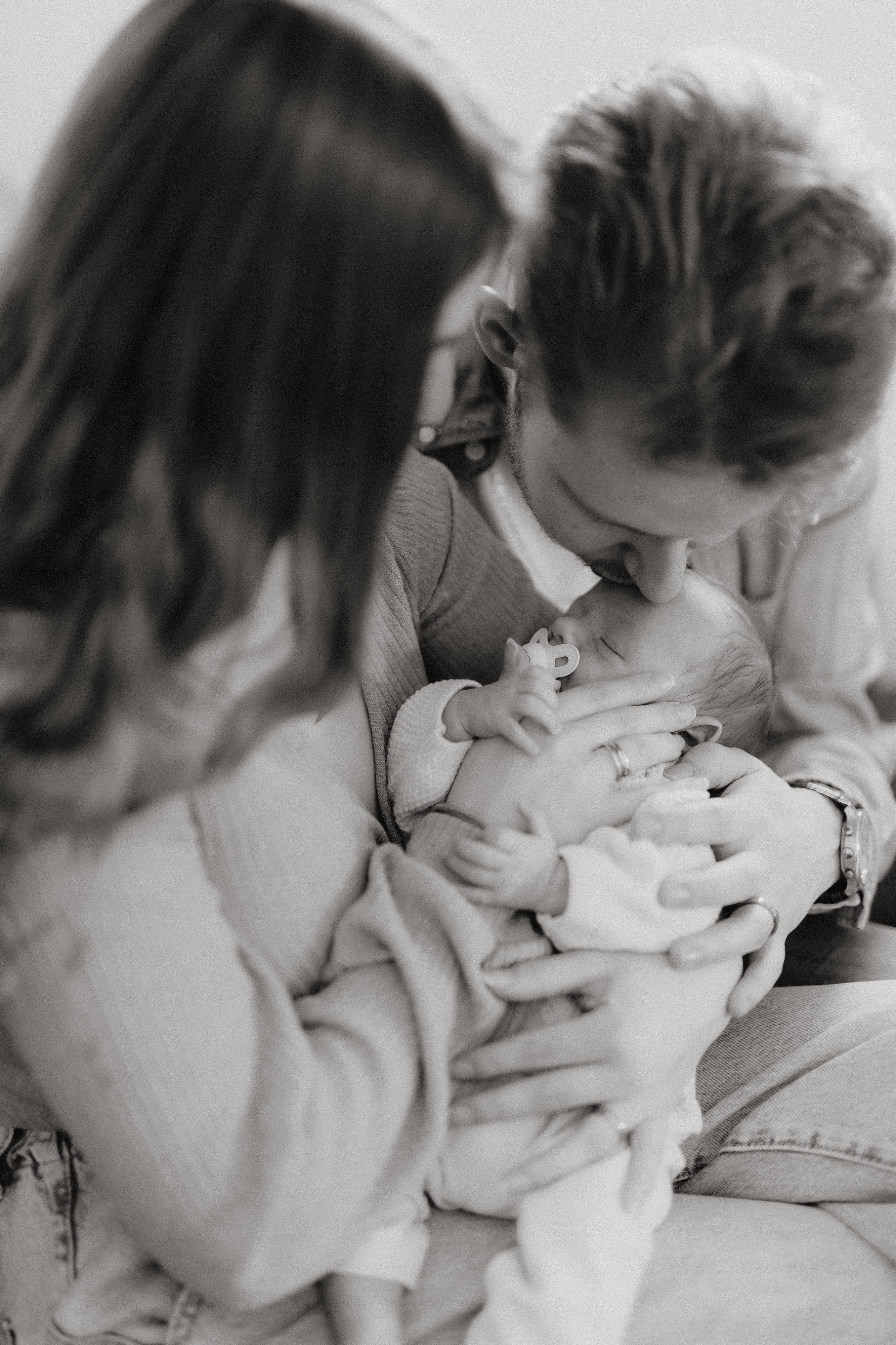 Schwarz-Weiß-Foto eines Paares, das sein Neugeborenes liebevoll im Arm hält und bewundert, aufgenommen während eines herzlichen Babyshootings. Sie sitzen eng beieinander, wobei der Vater das Baby sanft auf die Stirn küsst, während die Mutter liebevoll zusieht.