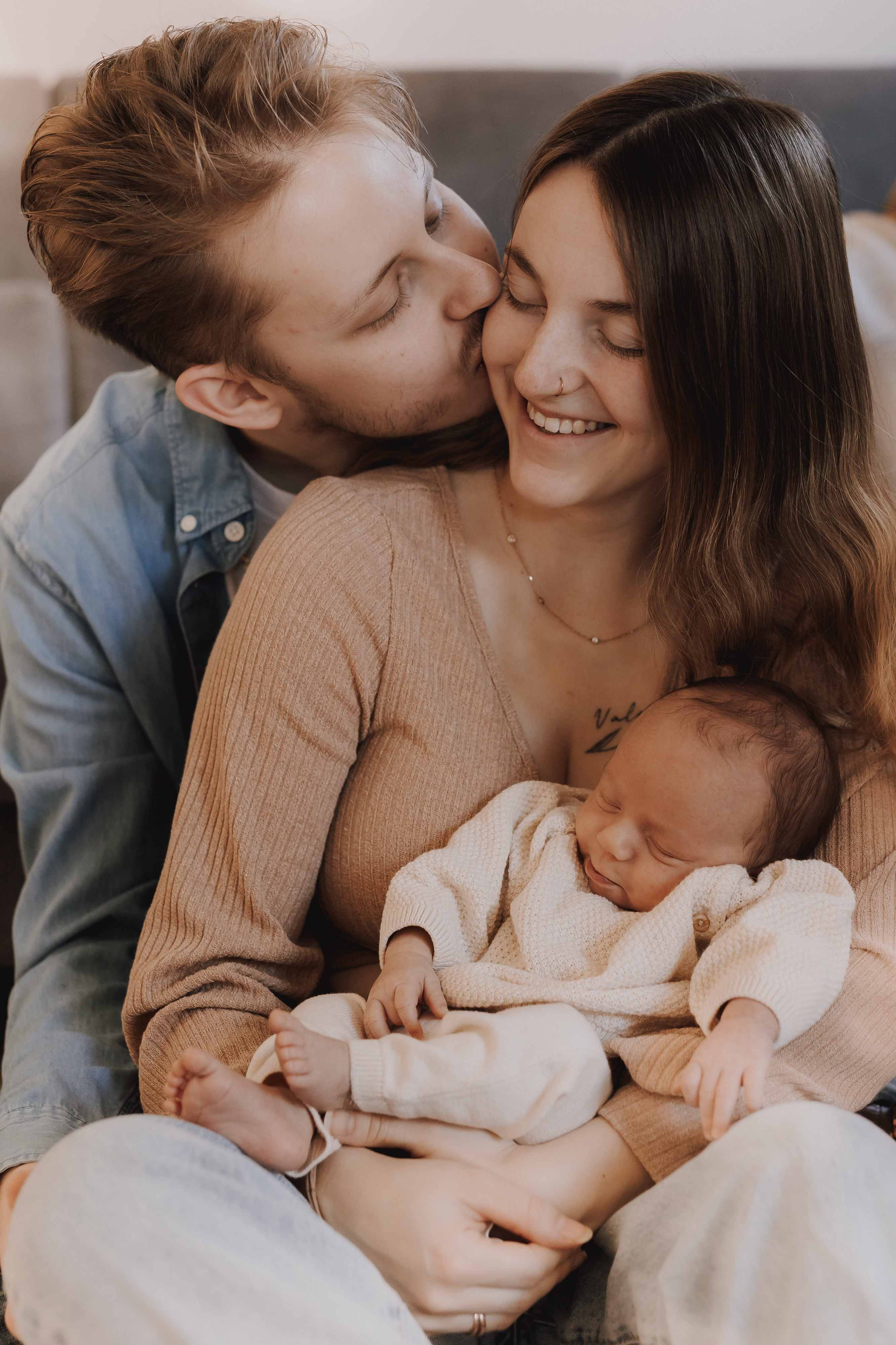 Ein Mann mit Bart küsst eine lächelnde Frau auf die Wange, während sie ein schlafendes Baby in den Armen hält und fängt damit die Essenz eines intimen Newbornshootings zu Hause ein. Die Frau trägt einen hellbraunen Pullover und das Baby ist in ein beiges Strickoutfit gekleidet, alles sitzt auf einem grauen Polstermöbel.