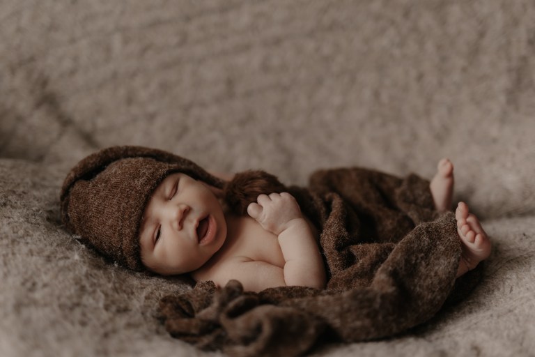Ein Neugeborenes schläft friedlich auf einer weichen, strukturierten Oberfläche während eines Fotoshootings von Tasha Ender Photography. Das Baby ist in eine braune, flauschige Decke gehüllt und trägt eine passende Strickmütze mit Schlappohren. Der Mund des Babys ist leicht geöffnet und eine Hand ist teilweise in der Nähe seines Gesichts gekrümmt.