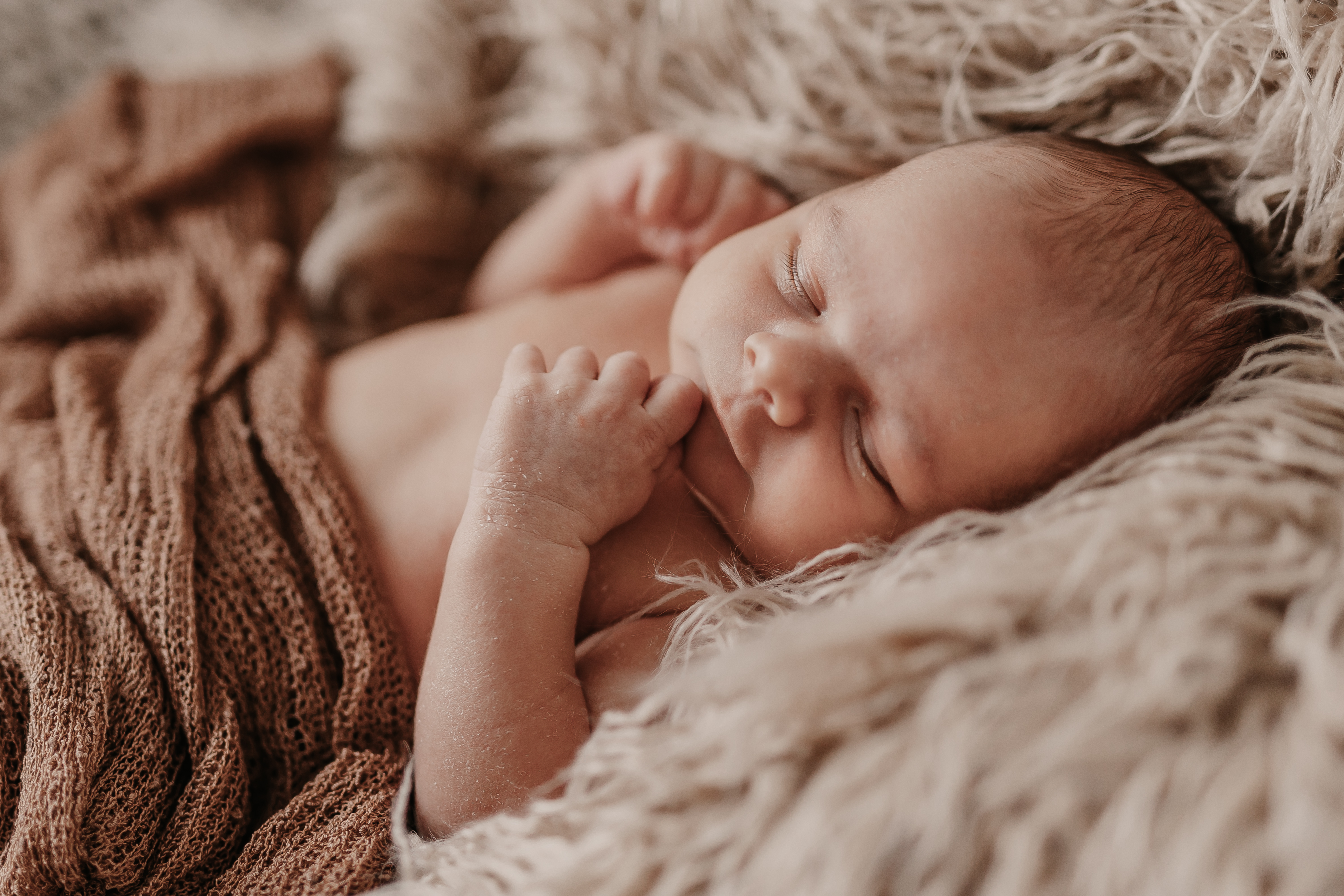 Ein schlafendes Neugeborenes ist in eine weiche, braune Decke gewickelt und liegt auf einer flauschigen, beigen Oberfläche. Das Baby hat die Augen geschlossen und die Hände ruhen sanft neben seinem Gesicht, was eine friedliche und ruhige Neugeborenenfotoszene ergibt, die von einer erfahrenen Neugeborenenfotografin aufgenommen wurde.
