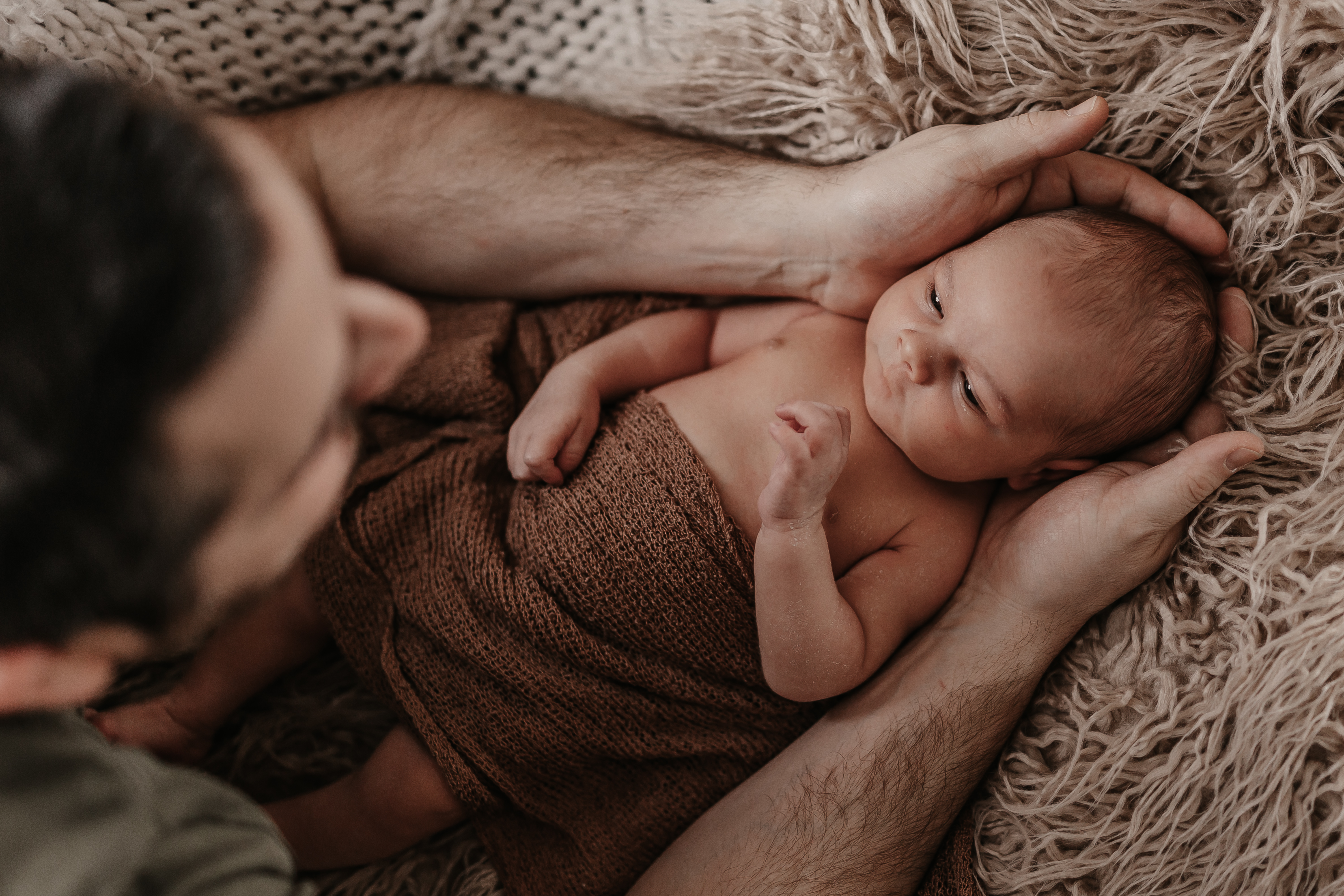 Ein Neugeborenes, in eine braune Strickdecke gehüllt, wird während eines Fotoshootings sanft von einem Erwachsenen, wahrscheinlich dem Elternteil, in den Armen gehalten. Das Baby liegt auf einer strukturierten, flauschigen Oberfläche und blickt nach oben. Die Szene, festgehalten von einer talentierten Neugeborenenfotografin in Baden, strahlt Wärme und Zärtlichkeit aus.