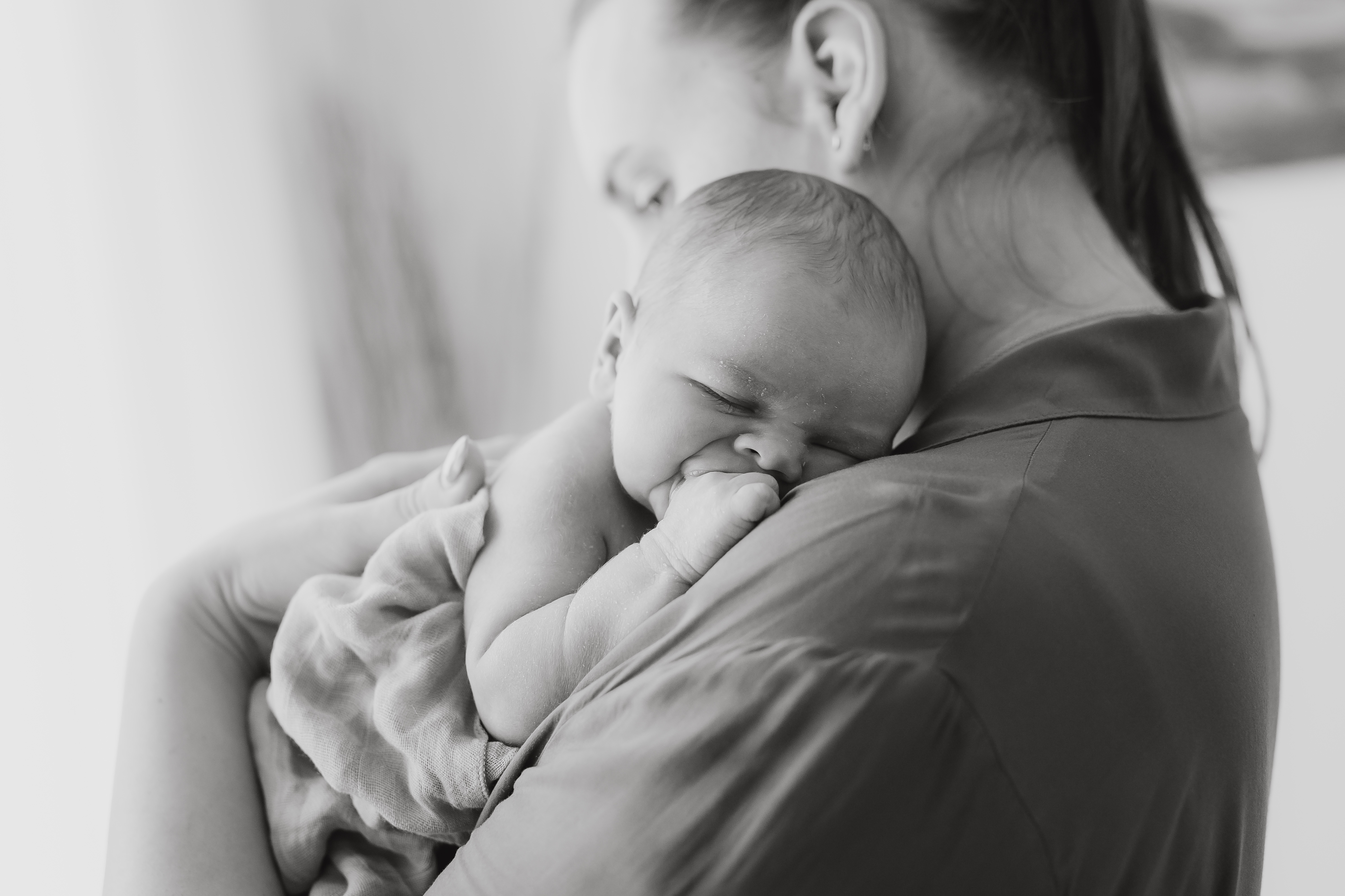 Ein Graustufenbild einer Person, die ein schlafendes Baby sanft an ihre Schulter hält. Der Kopf des Babys ruht auf der Schulter der Person, die Hand des Babys liegt nahe seinem Gesicht und die Augen sind geschlossen, was ein Gefühl der Ruhe und Geborgenheit vermittelt, das für Neugeborenenfotos charakteristisch ist.